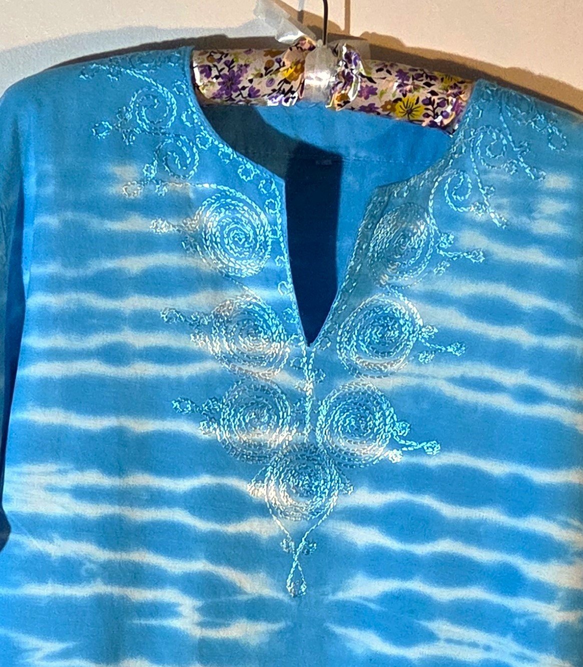 clode up of top of A light blue cotton kaftan shirt with embroidered details, showcasing a vertical tie-dye pattern of thin, wire-like white ripple lines.