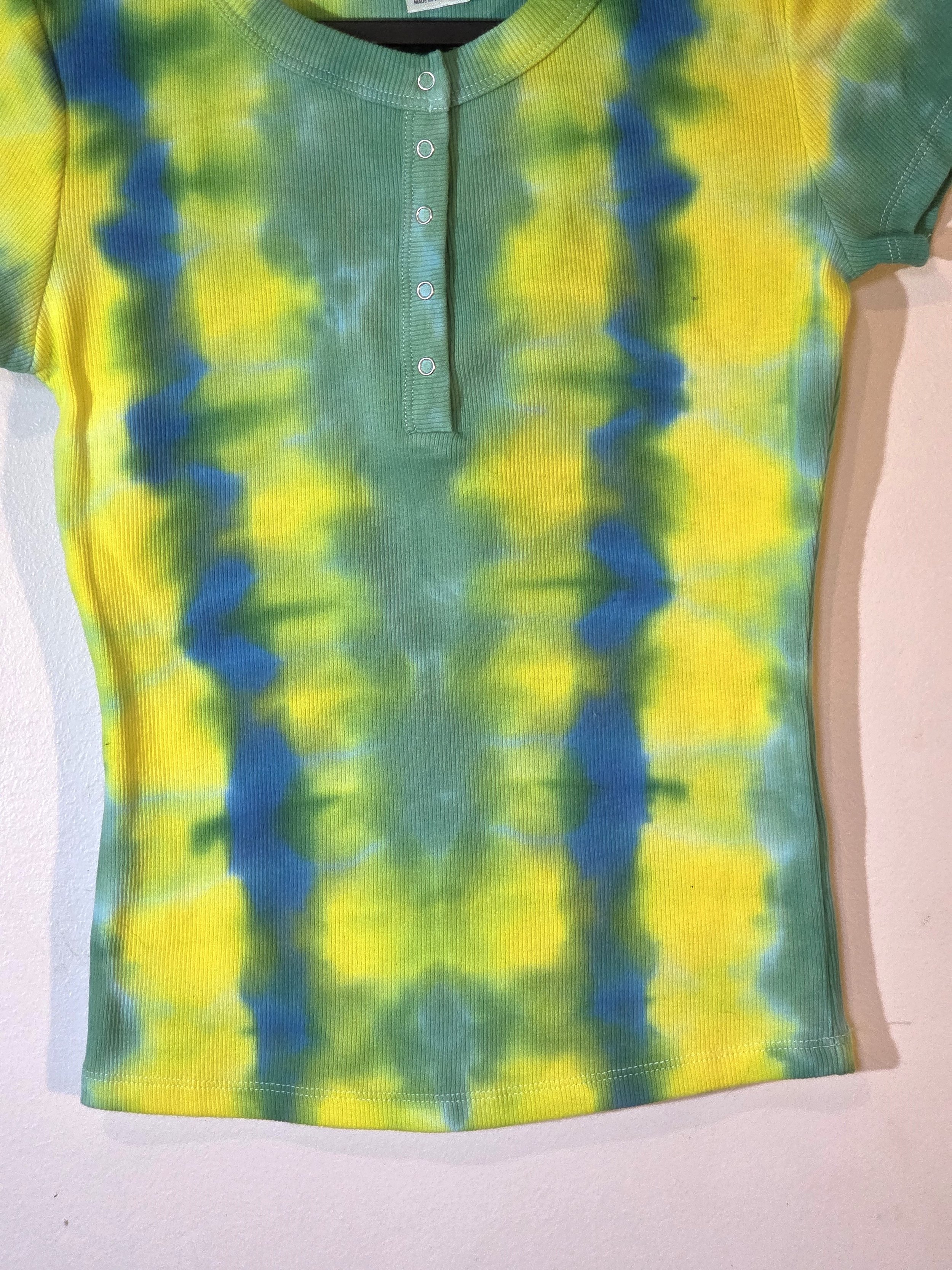 A child's ribbed t-shirt with front press studs, showcasing a vertical tie-dye pattern of alternating yellow, green, and blue-green columns.