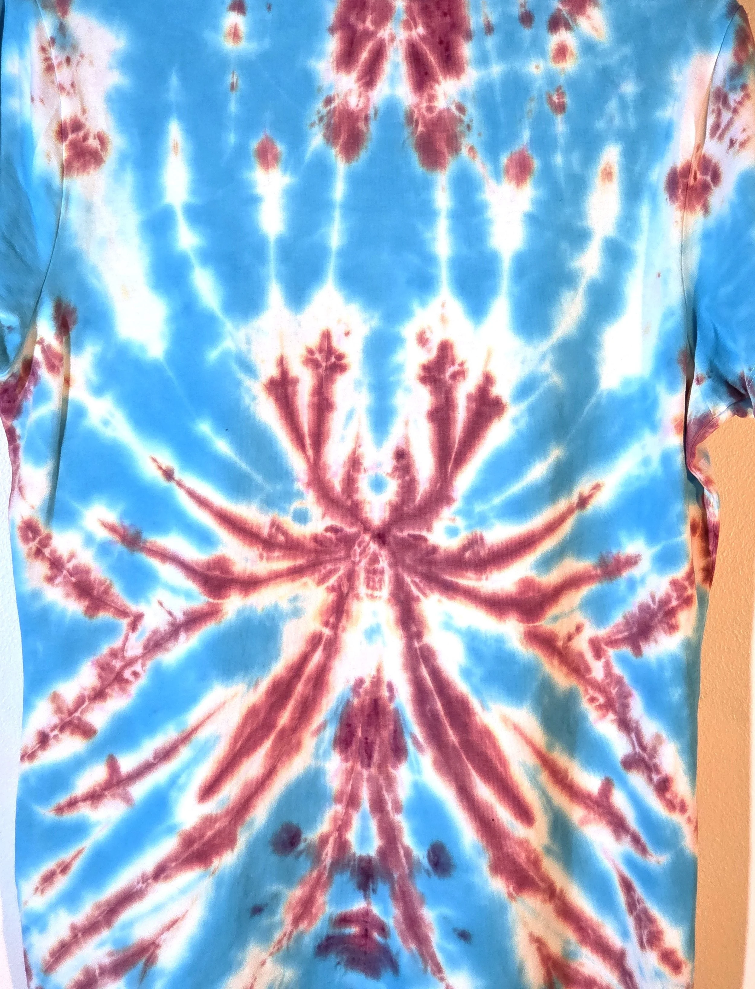back view of A size 10 t-shirt featuring a tie-dye pattern of a sharp, spiky purple and wine flower on a sky blue and white background.