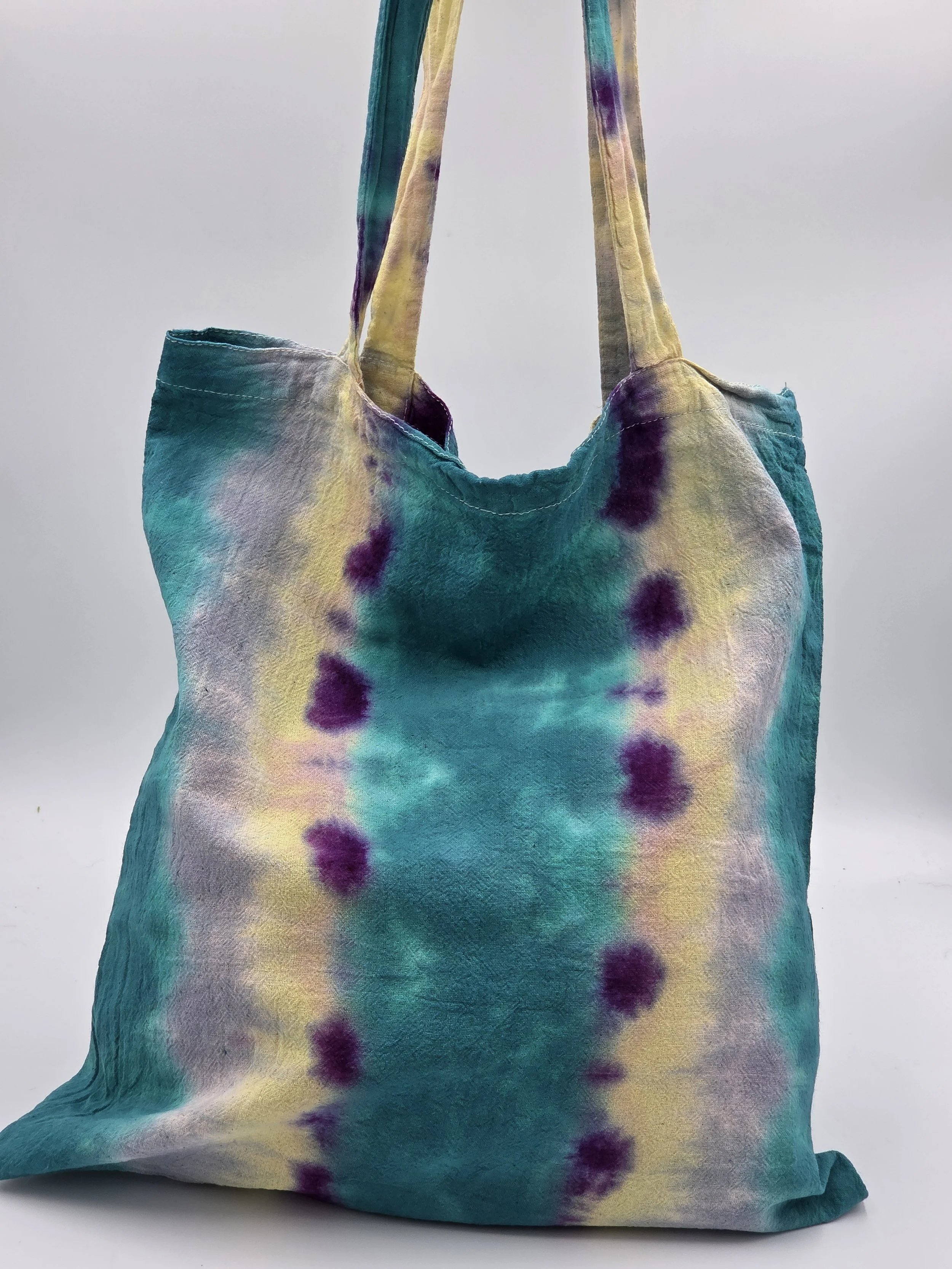 A calico tote bag featuring vertical tie-dye stripes of teal, yellow, and lavender with purple splotches along the center.