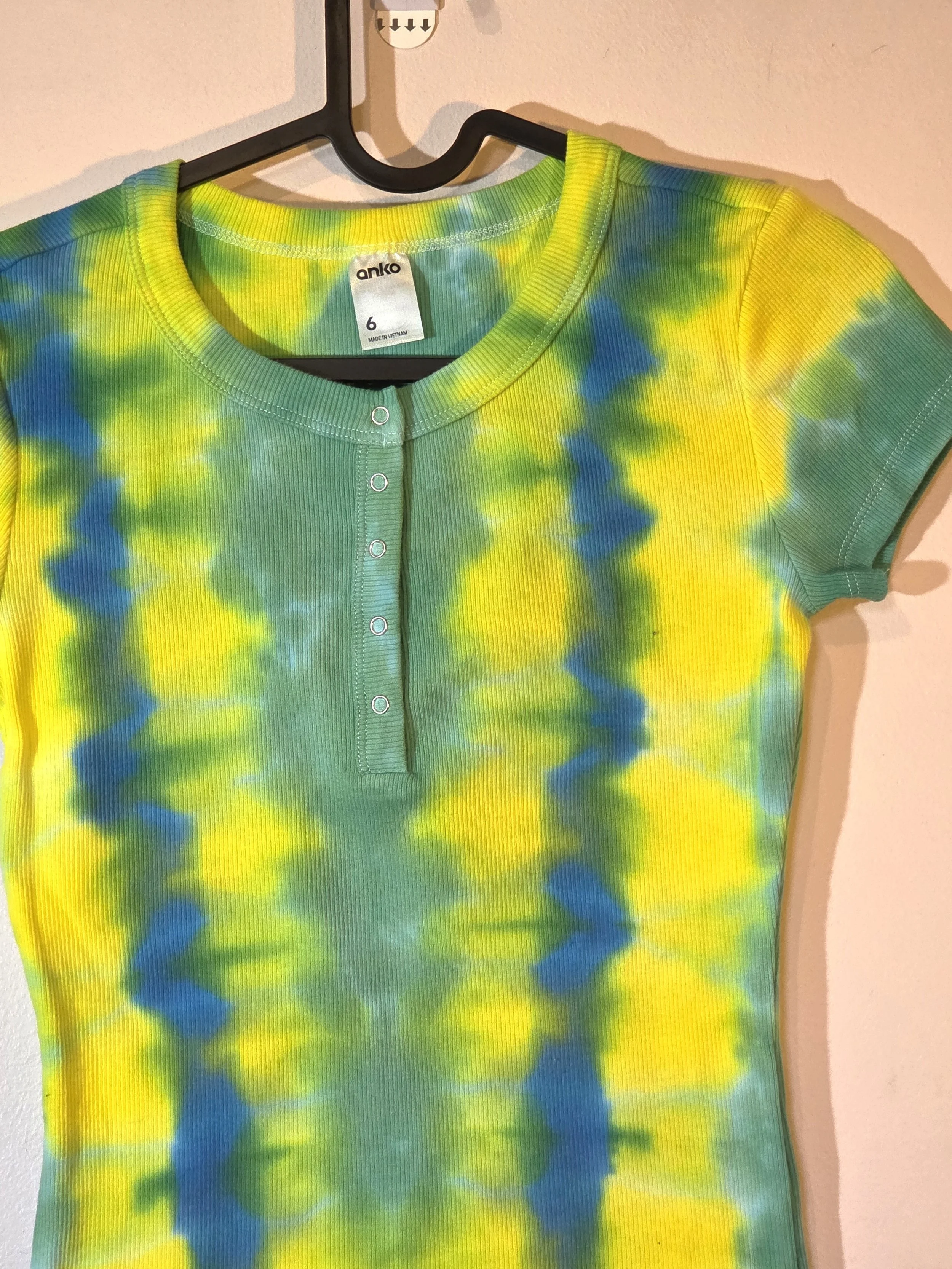 A child's ribbed t-shirt with front press studs, showcasing a vertical tie-dye pattern of alternating yellow, green, and blue-green columns.