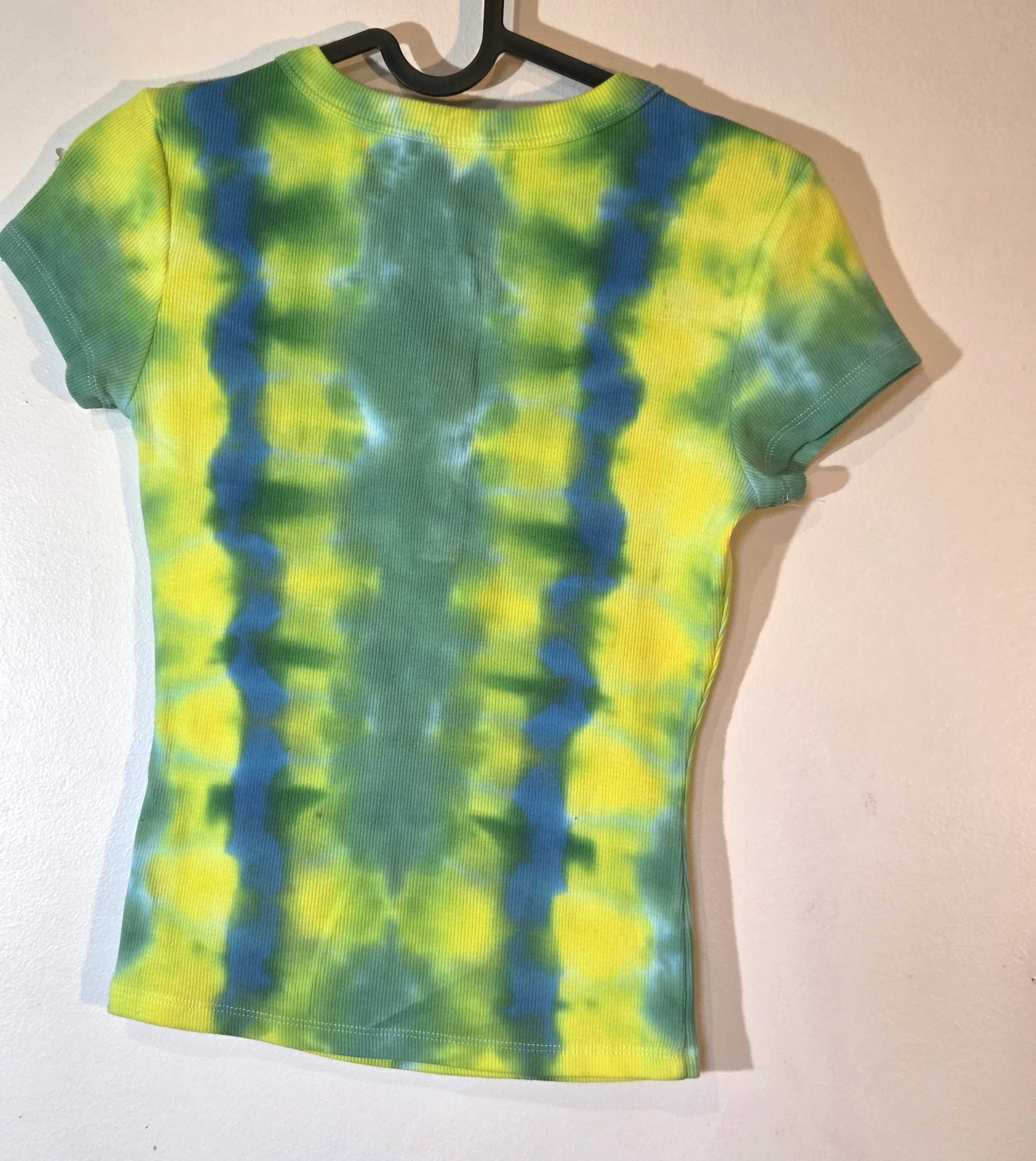 back view of A child's ribbed t-shirt with front press studs, showcasing a vertical tie-dye pattern of alternating yellow, green, and blue-green columns.