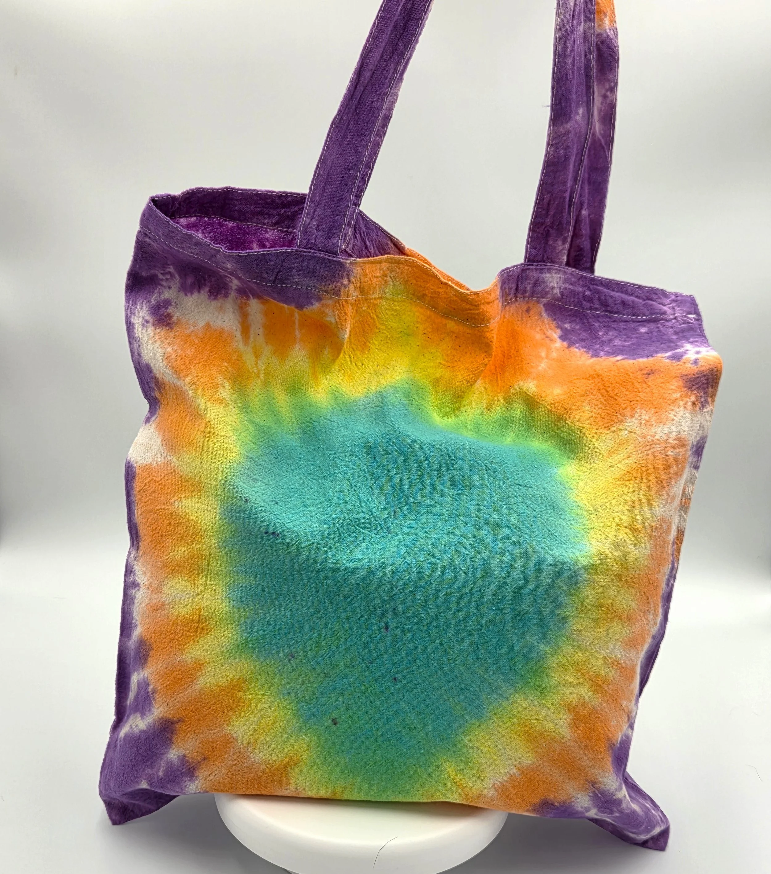 A calico tote bag featuring a tie-dye pattern with a large teal oval center surrounded by glowing rings of yellow, orange, and purple.