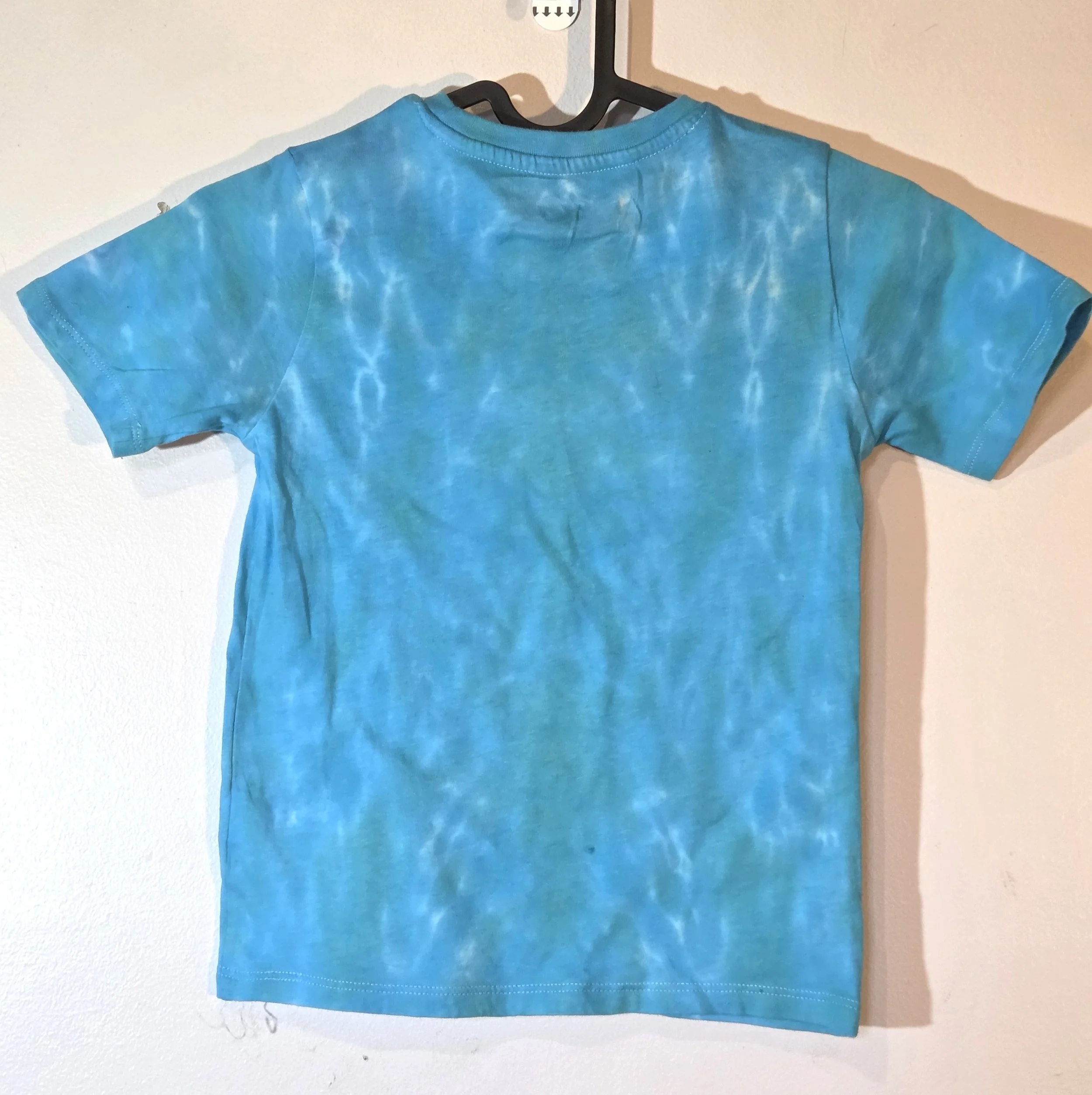 back view of A child's sky blue t-shirt displaying a delicate, flowing tiger stripe tie-dye pattern with thin white veins near the shoulders.