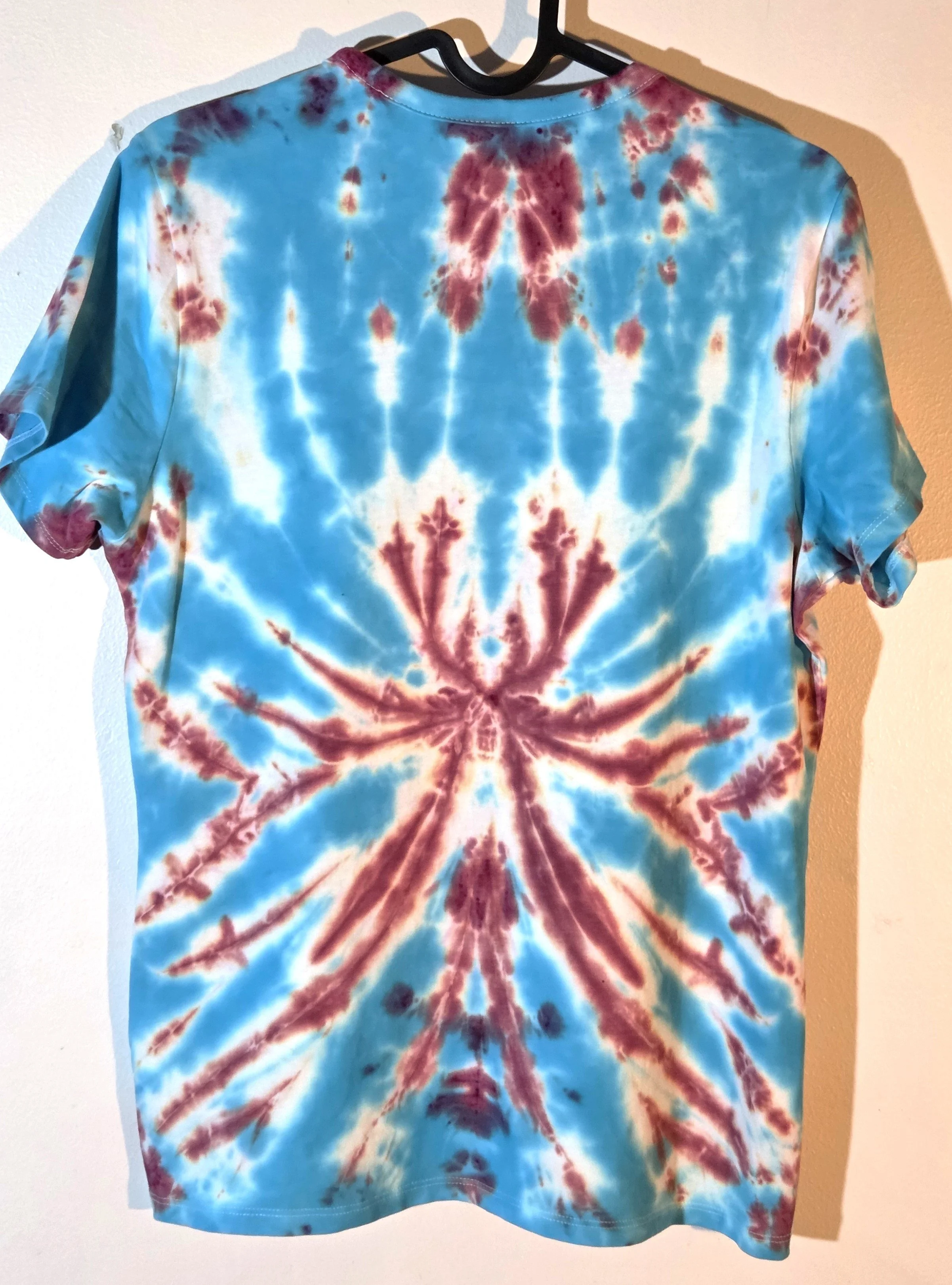 Back view of A size 10 t-shirt featuring a tie-dye pattern of a sharp, spiky purple and wine flower on a sky blue and white background.