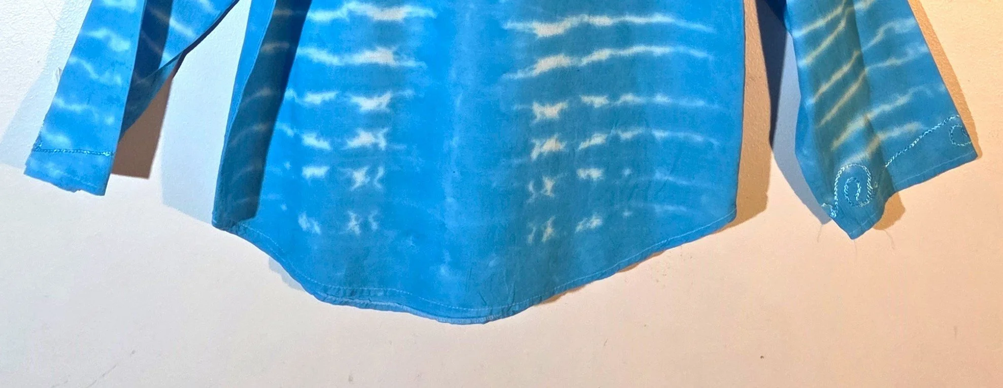 back hem of A light blue cotton kaftan shirt with embroidered details, showcasing a vertical tie-dye pattern of thin, wire-like white ripple lines.