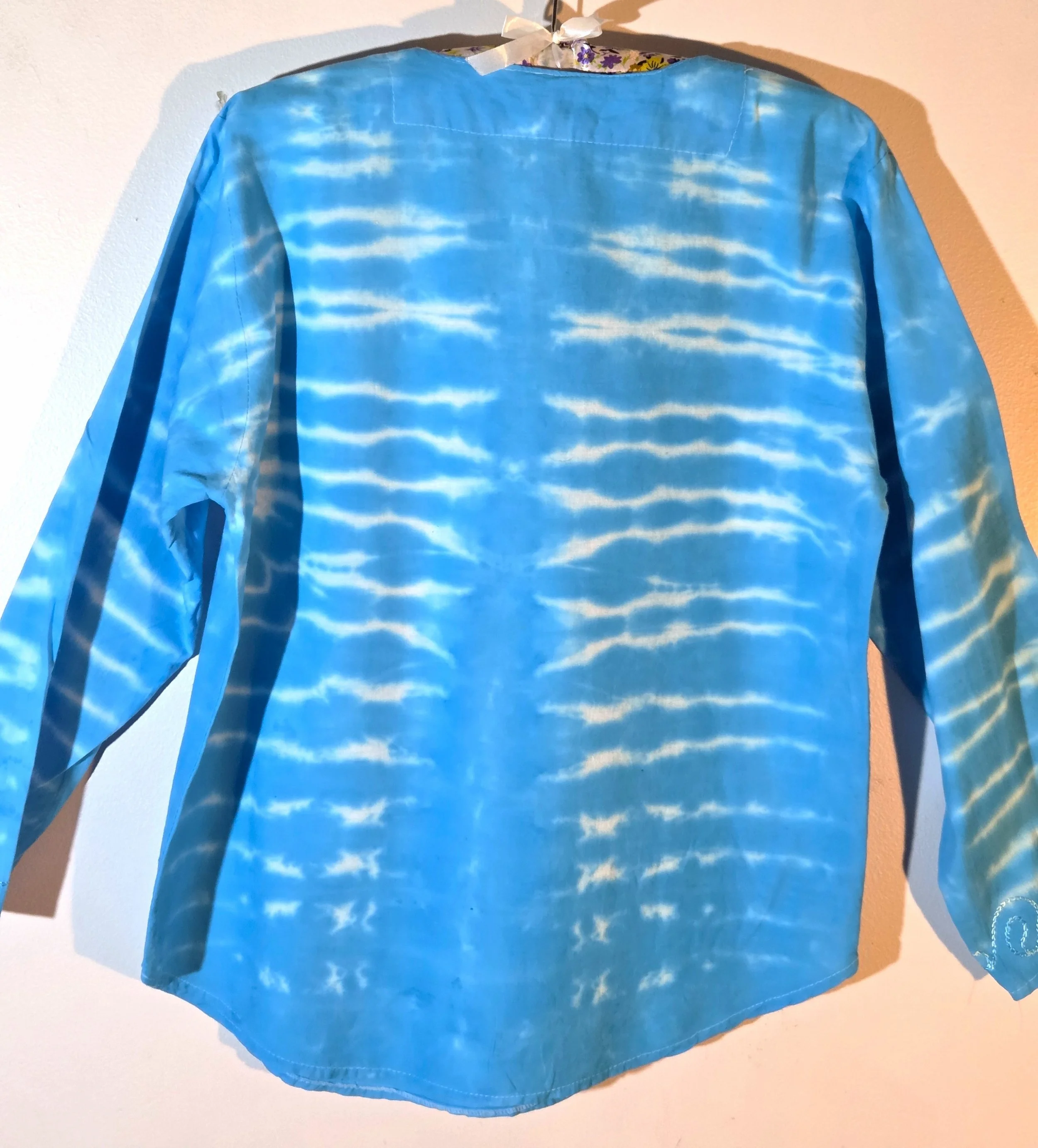back view of A light blue cotton kaftan shirt with embroidered details, showcasing a vertical tie-dye pattern of thin, wire-like white ripple lines.