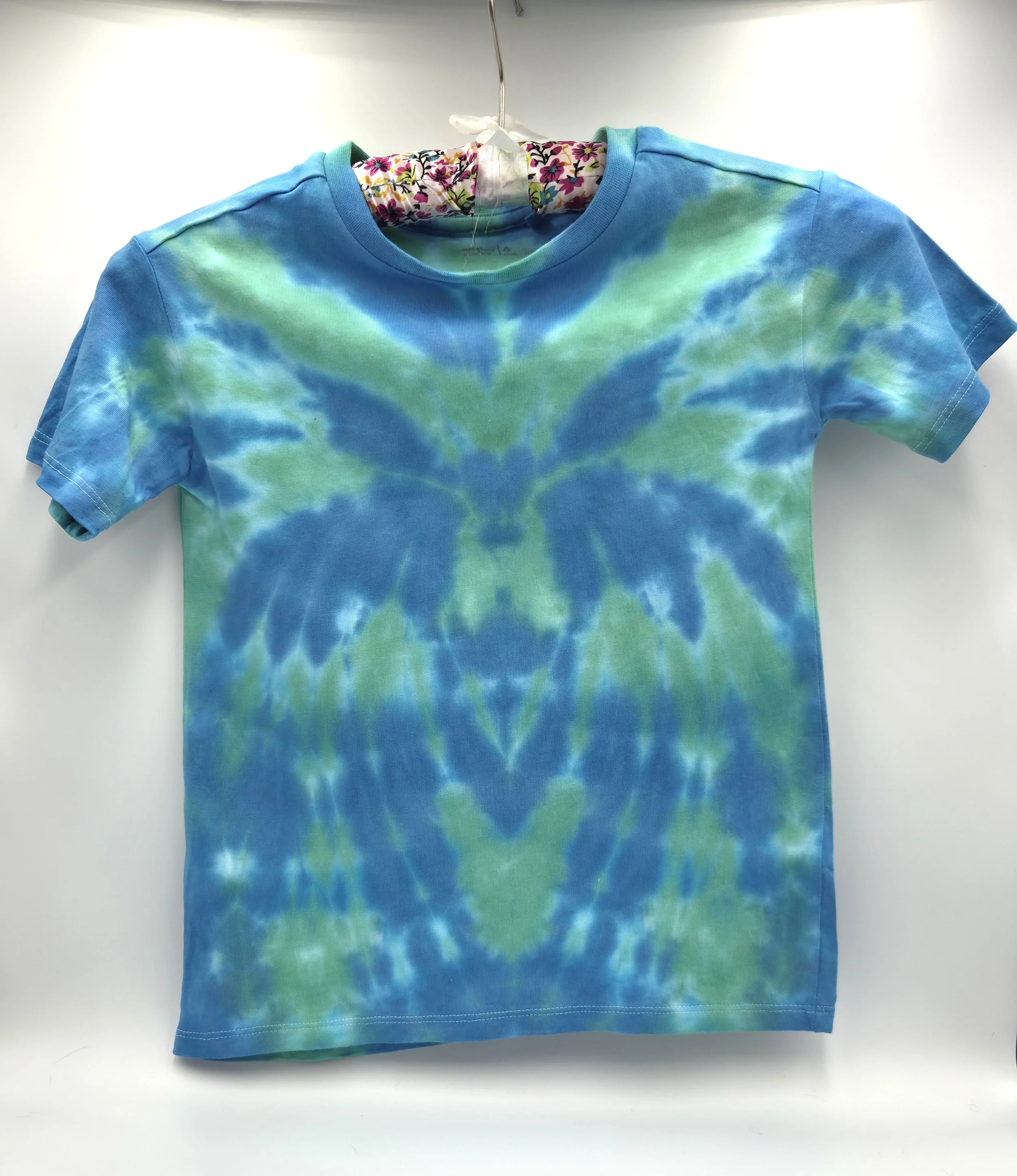 A child's light green t-shirt displaying a central sky blue tie-dye spider pattern, back view