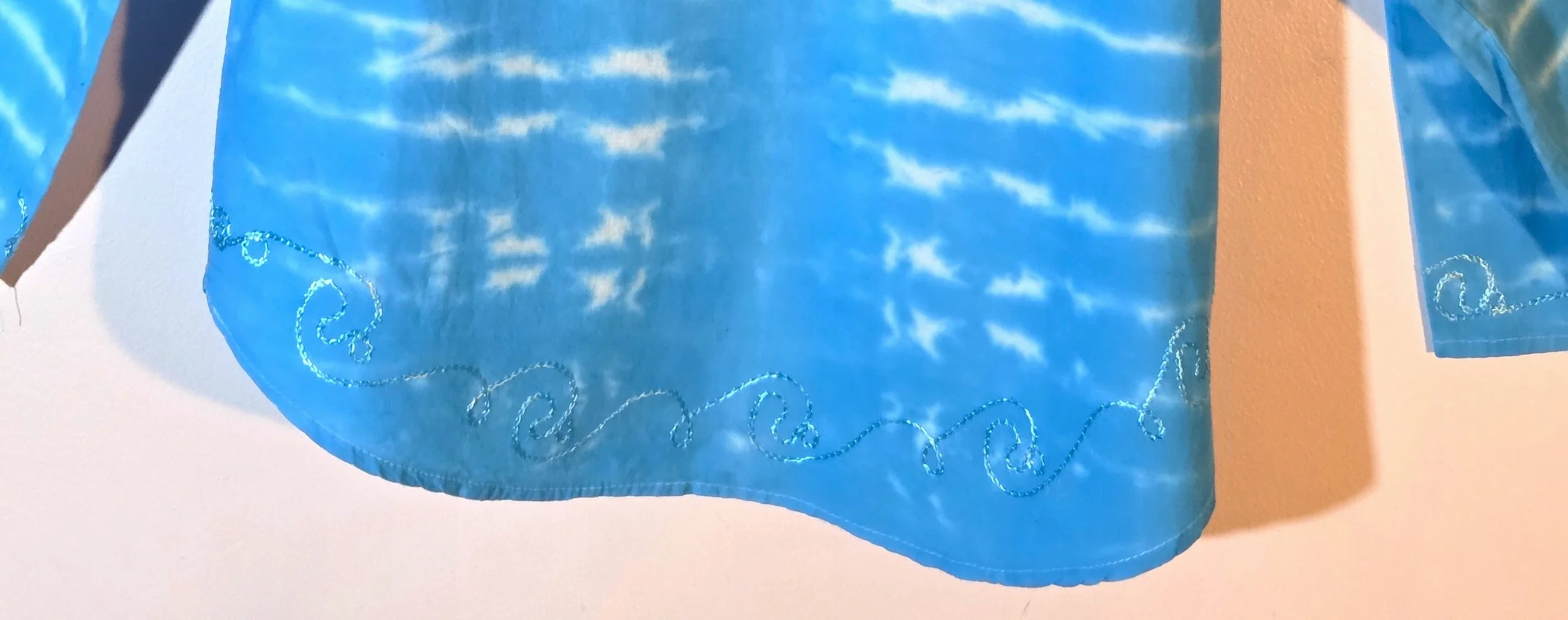 bottom hem of A light blue cotton kaftan shirt with embroidered details, showcasing a vertical tie-dye pattern of thin, wire-like white ripple lines.