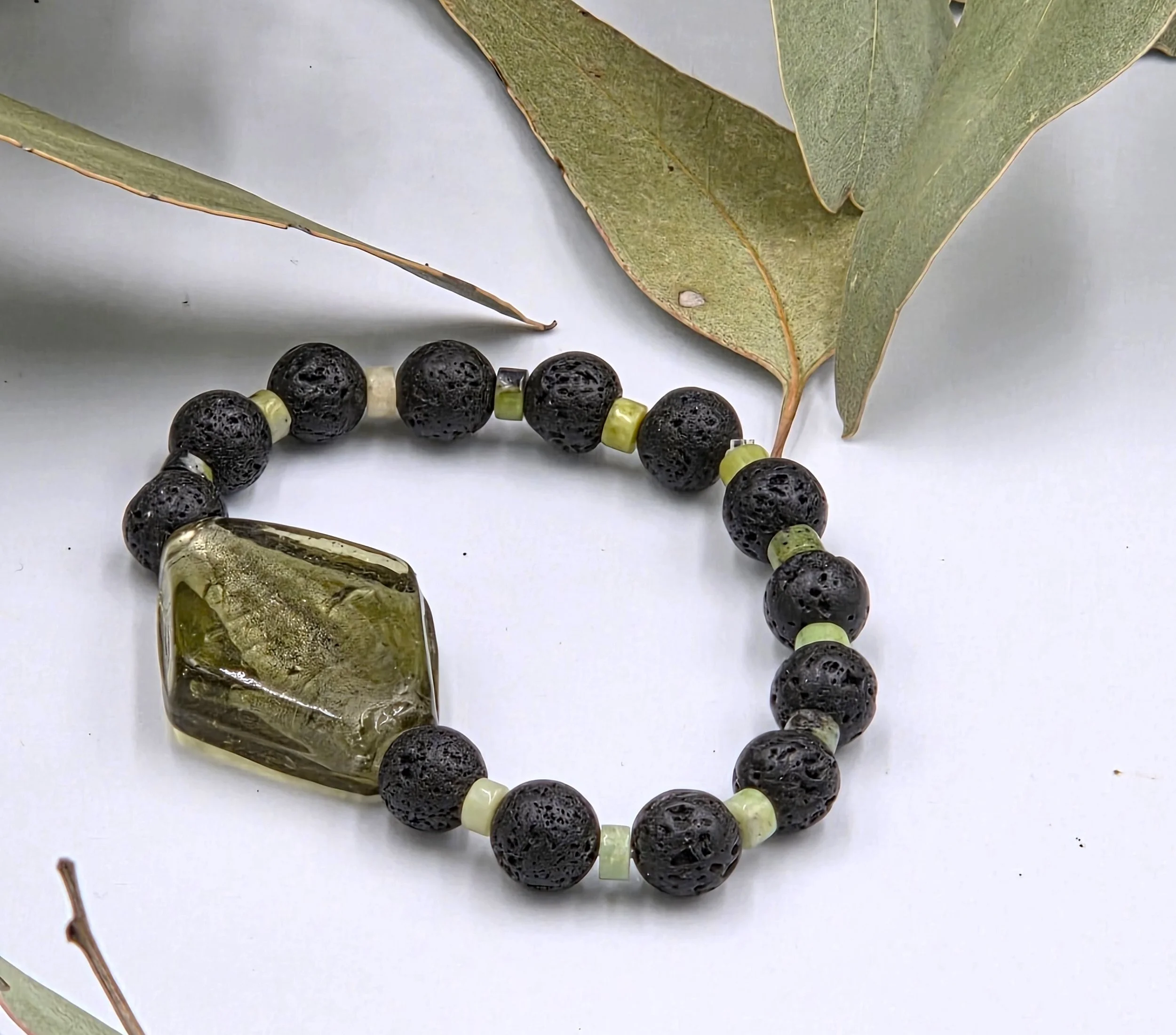 Olive green rhombus glass bead centered between alternating black lava beads and small green chrysoprase discs on an elastic bracelet.