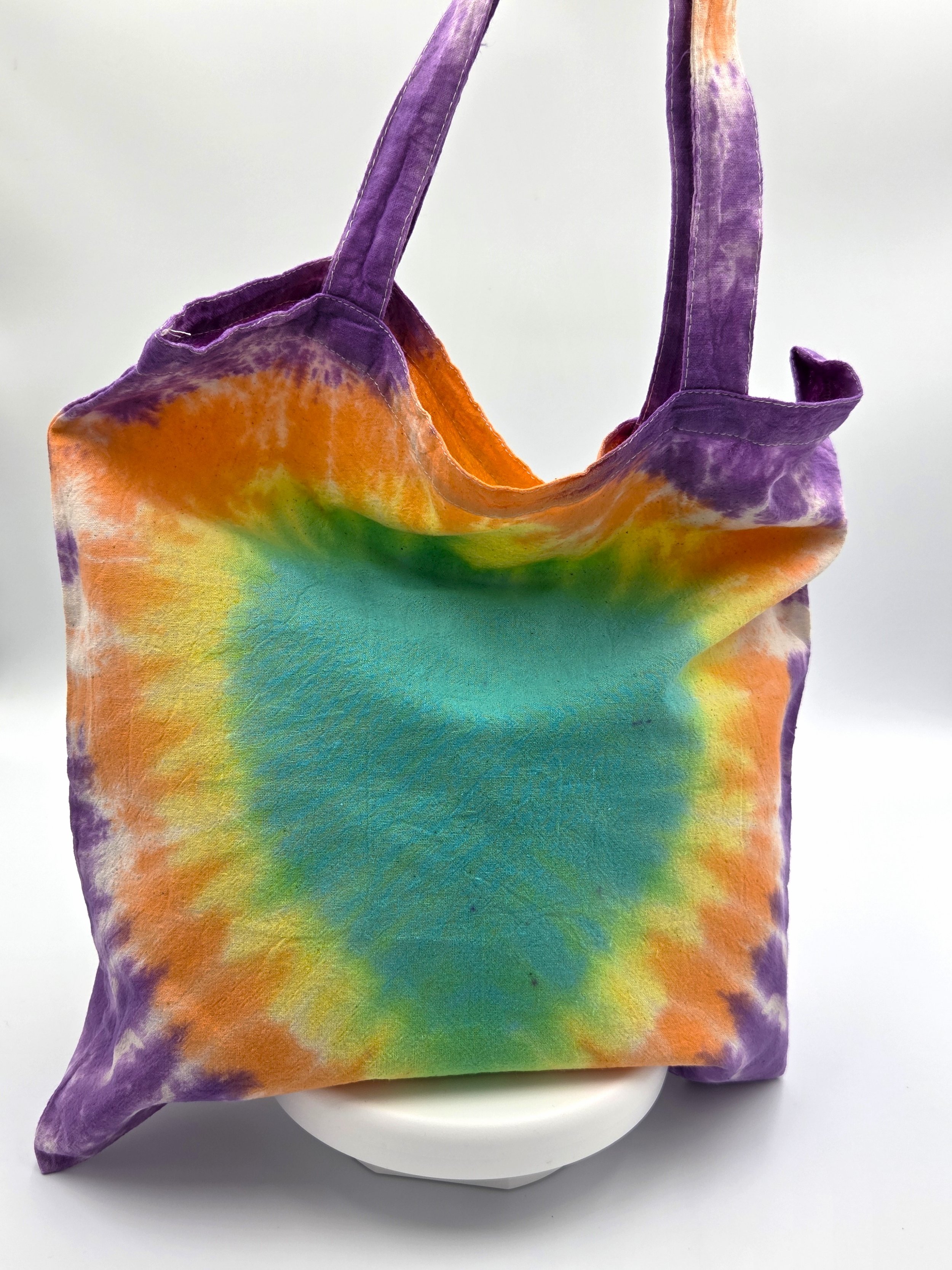 A calico tote bag featuring a tie-dye pattern with a large teal oval center surrounded by glowing rings of yellow, orange, and purple.