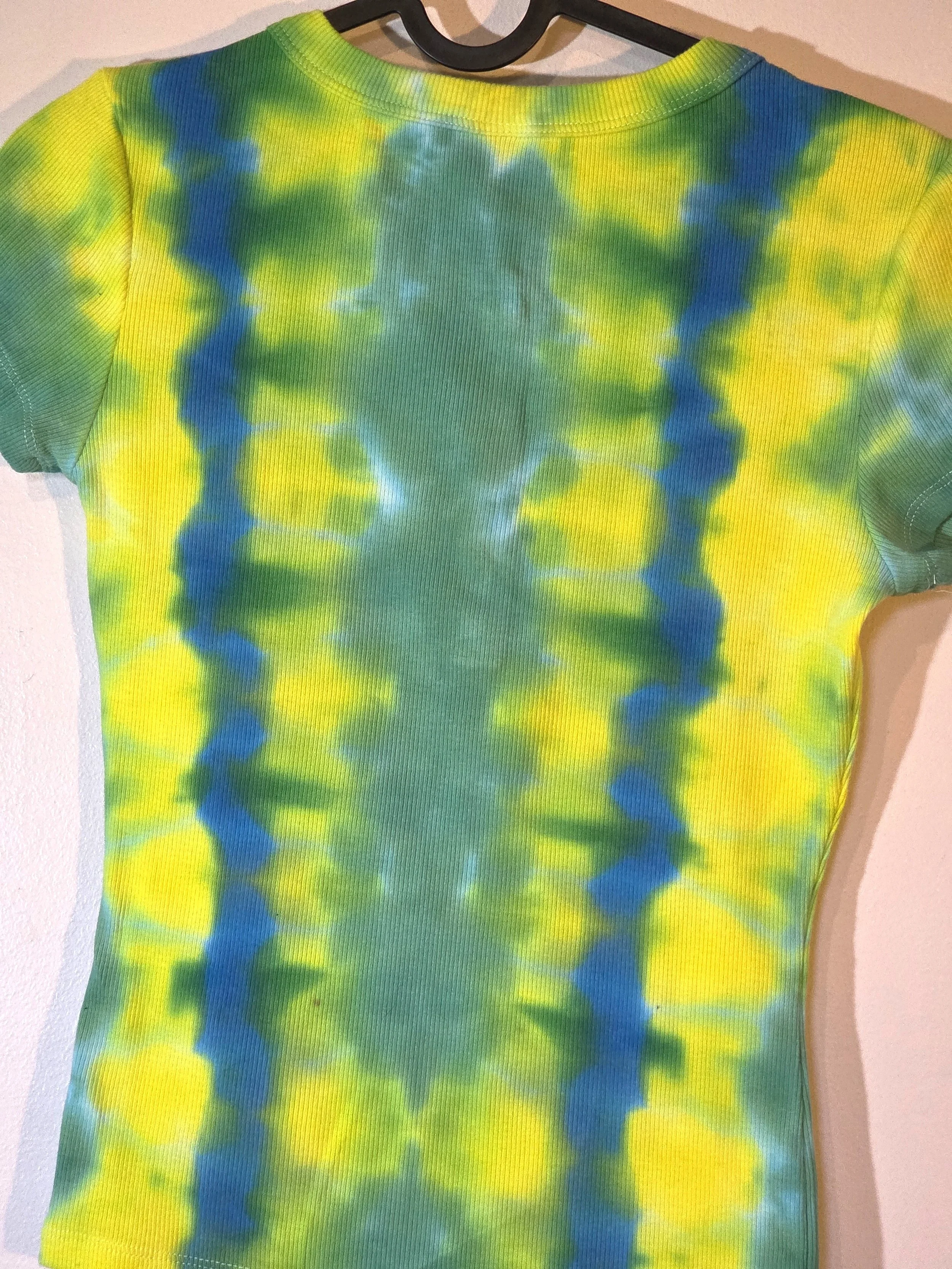 close up back view of A child's ribbed t-shirt with front press studs, showcasing a vertical tie-dye pattern of alternating yellow, green, and blue-green columns.