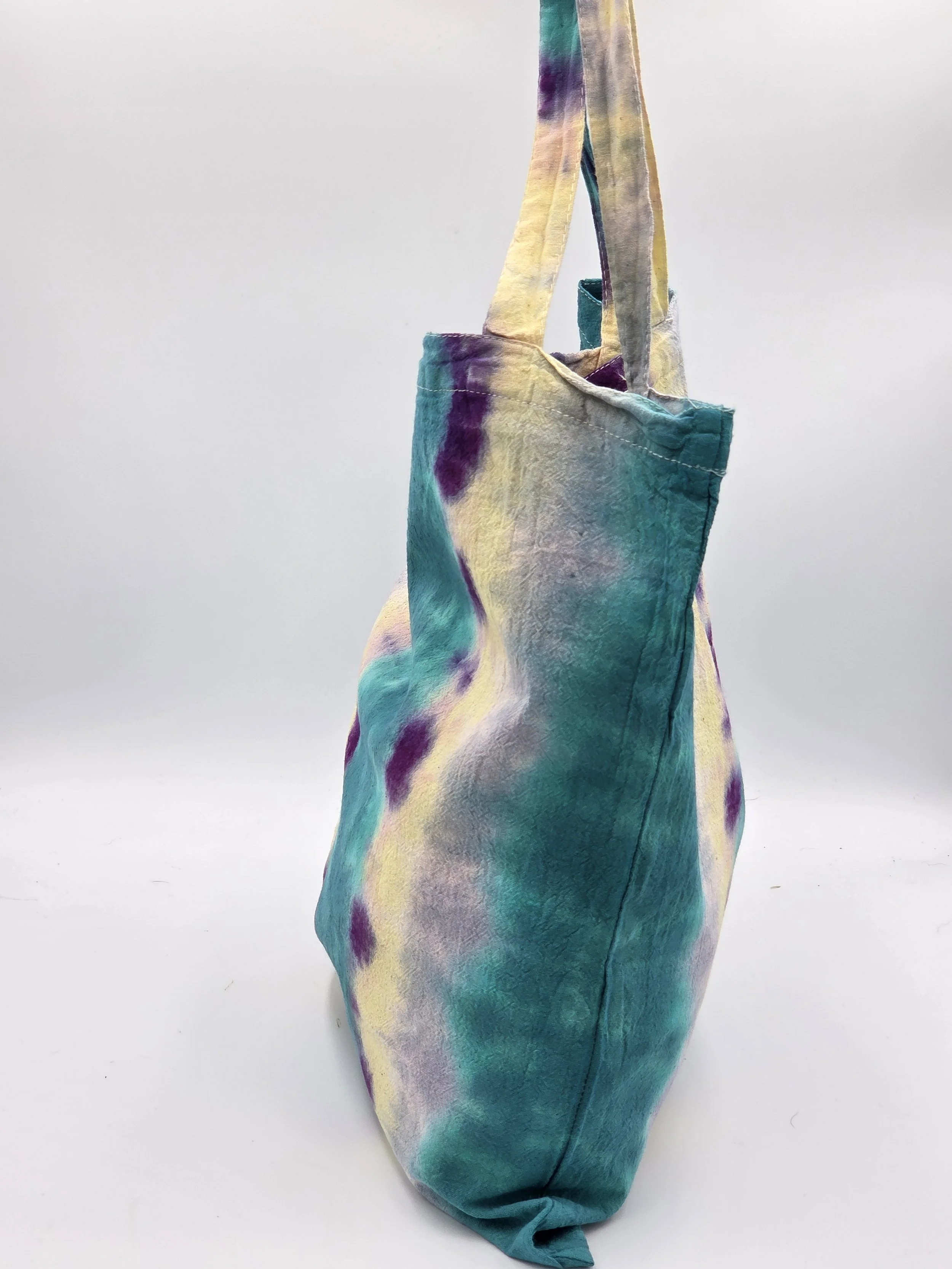 A calico tote bag featuring vertical tie-dye stripes of teal, yellow, and lavender with purple splotches along the center.