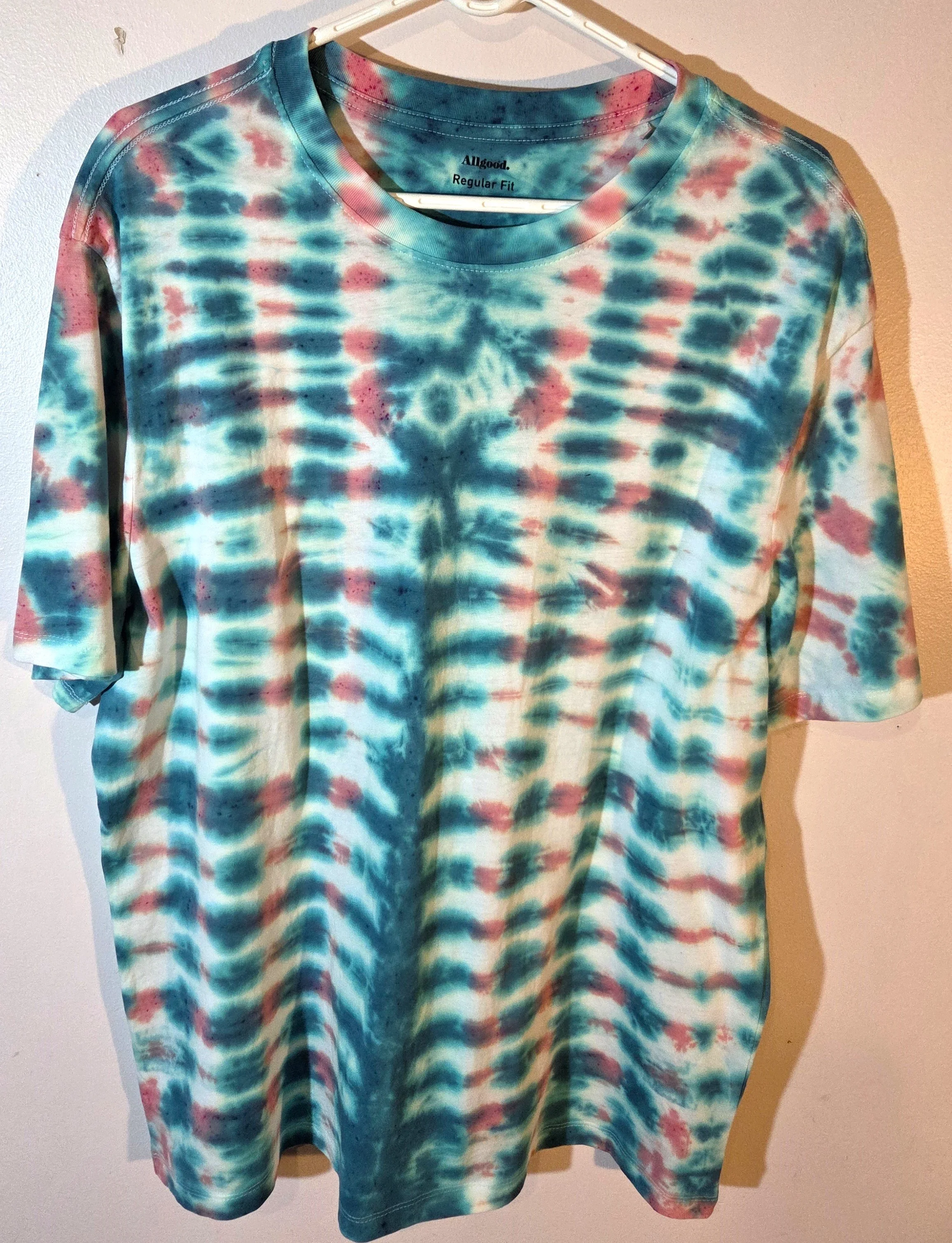 An XL adult white t-shirt featuring a tie-dye grid of Aegean green and coral with a prominent green centerline and eye motif near the neck.