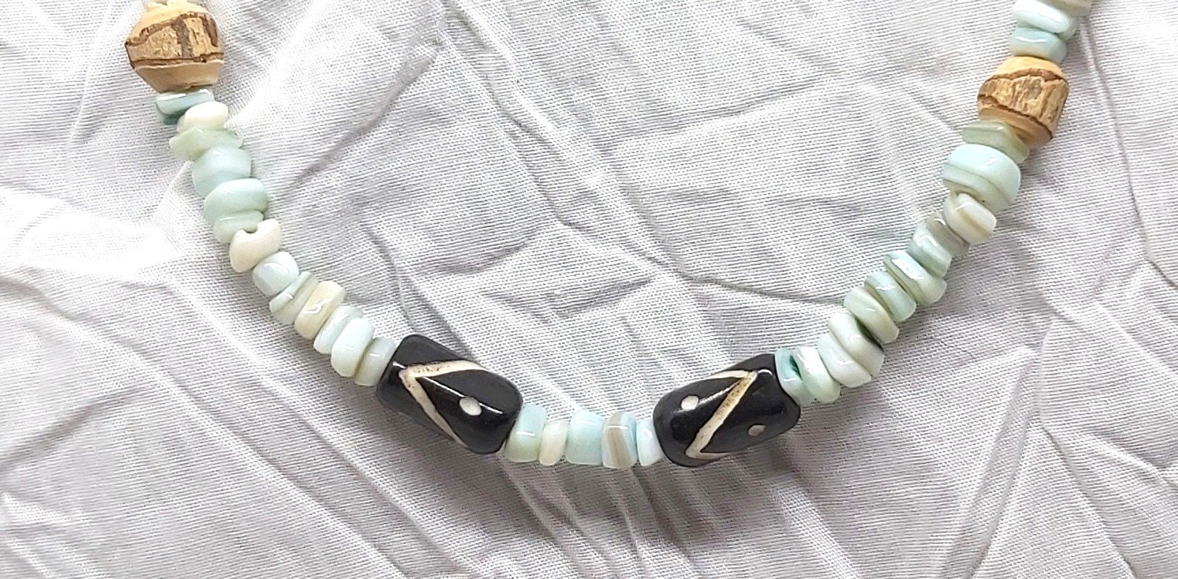 close up A necklace that has shell chips dyed blue-green and natural wood beads that have their original rough bark. The focal point is two dark brown acrylic carved tubes.