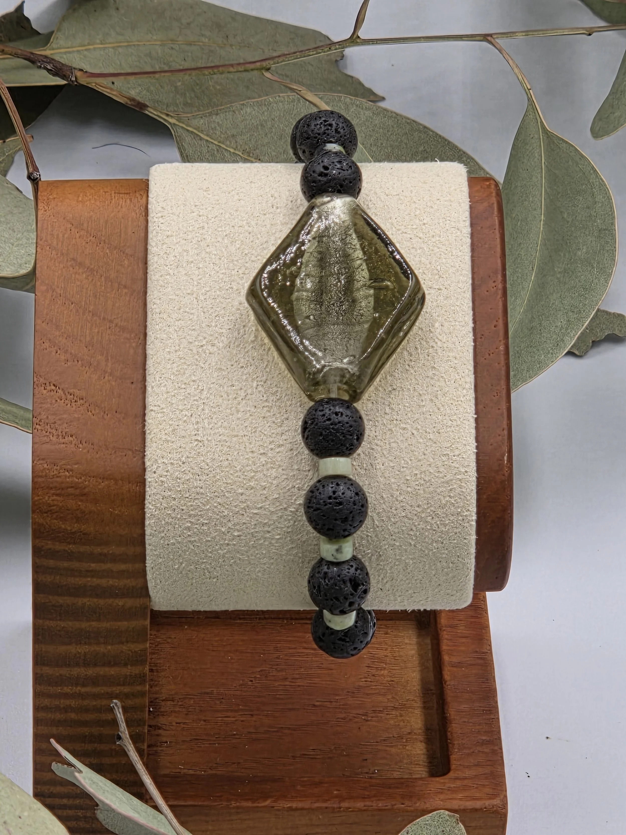 Olive green rhombus glass bead centered between alternating black lava beads and small green chrysoprase discs on an elastic bracelet.