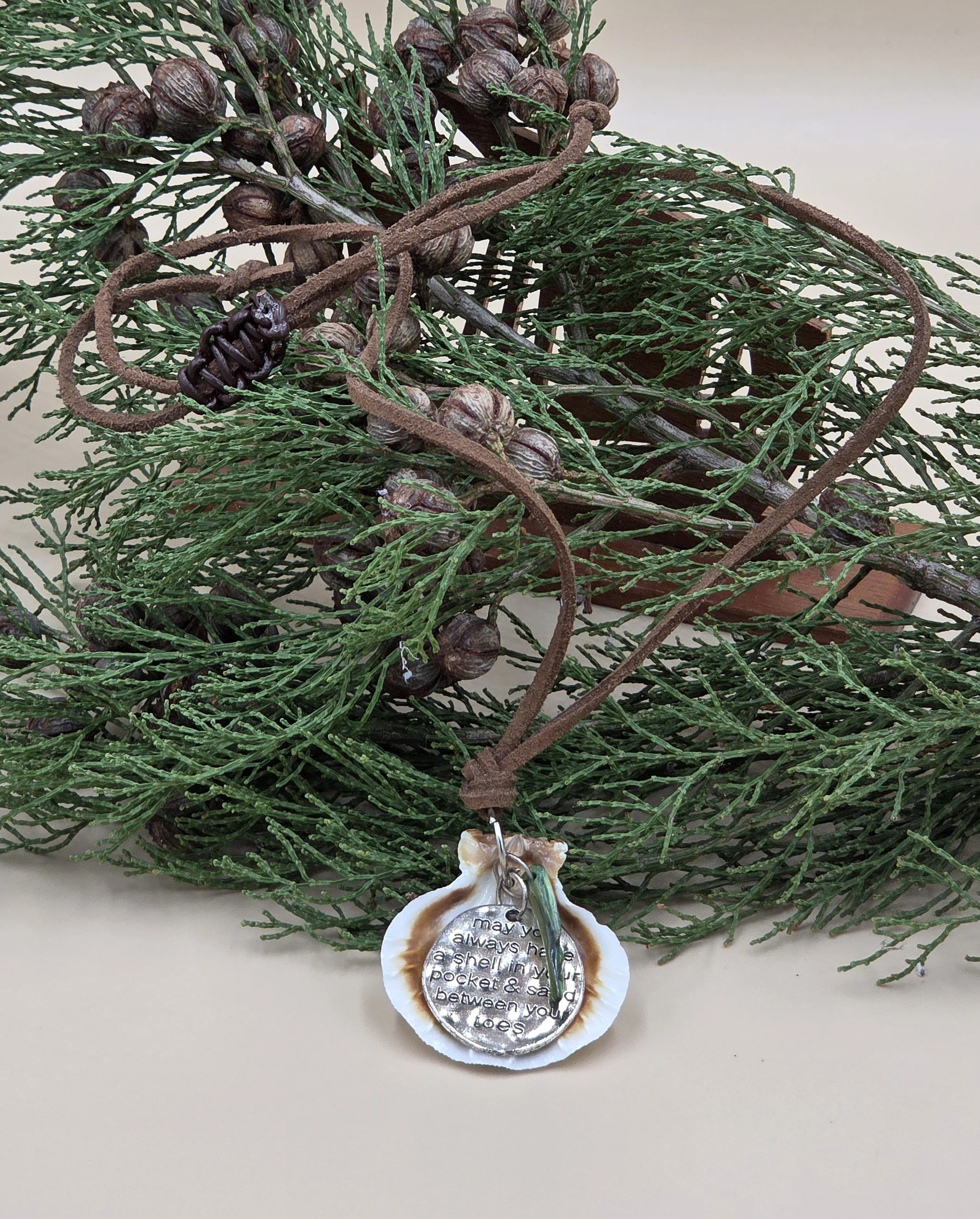 On background of native pine and nuts Australian handmade necklace with inscribed metal pendant and blue shell on suedette cord.