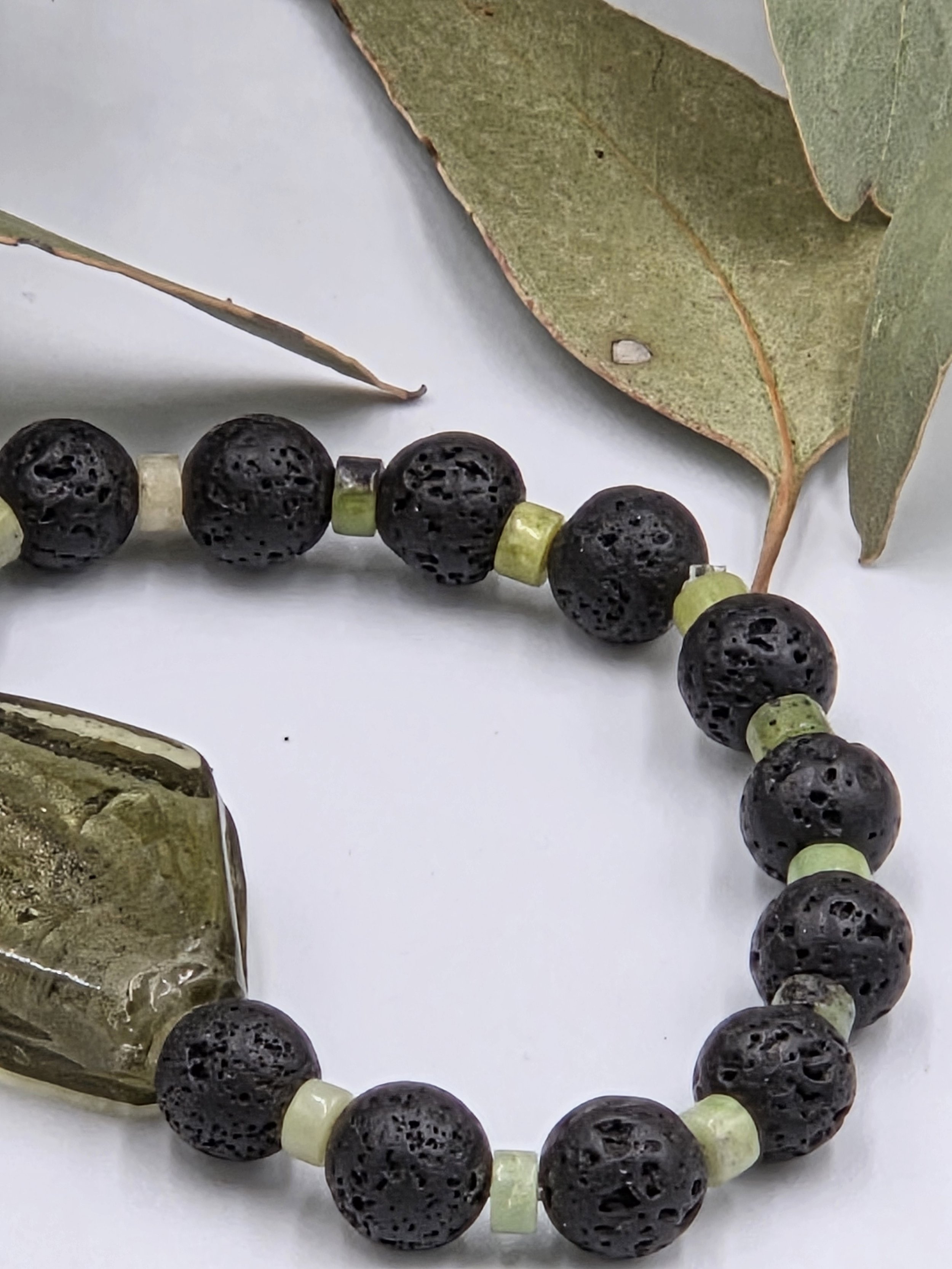 Olive green rhombus glass bead centered between alternating black lava beads and small green chrysoprase discs on an elastic bracelet.