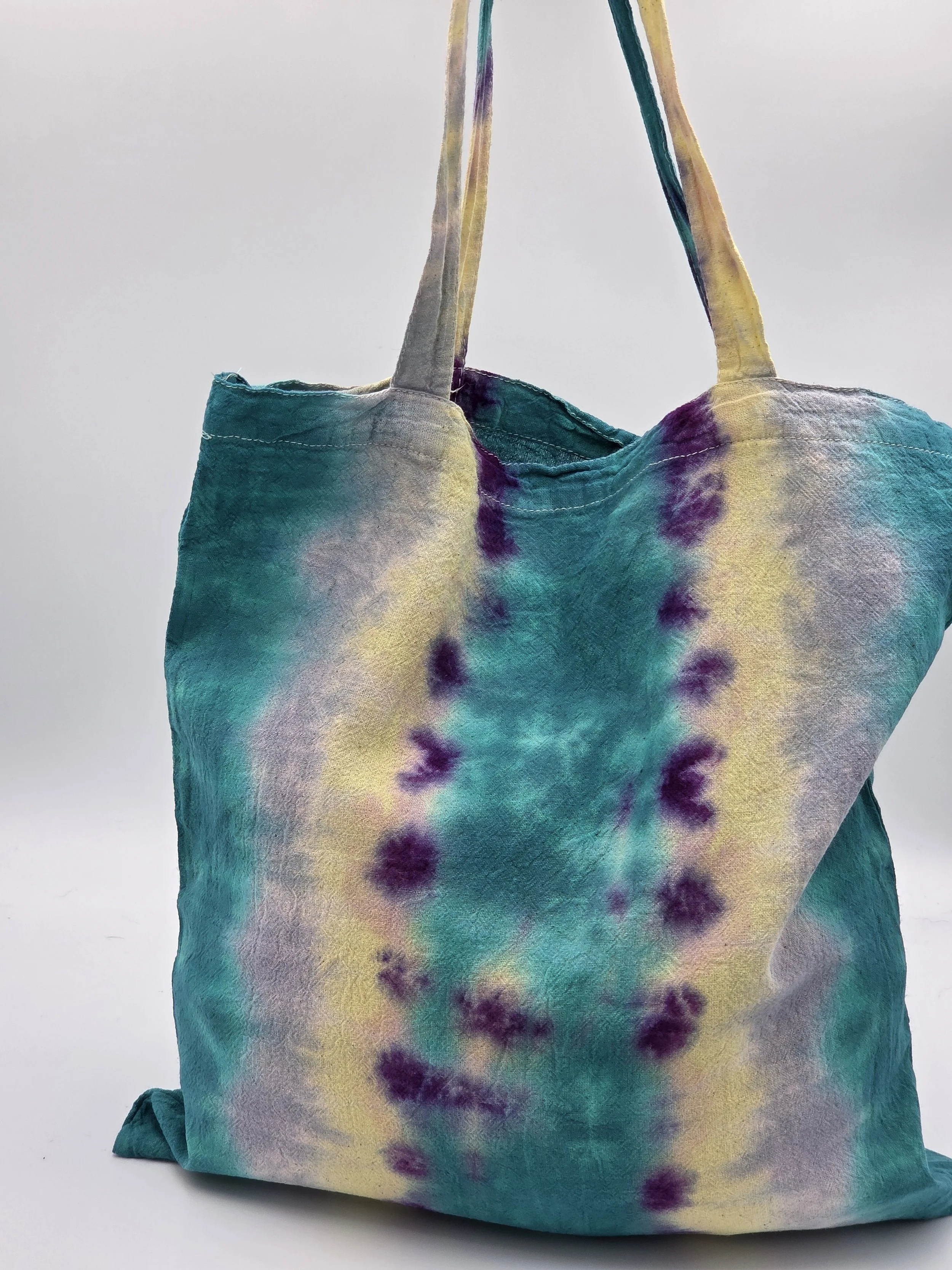 A calico tote bag featuring vertical tie-dye stripes of teal, yellow, and lavender with purple splotches along the center.