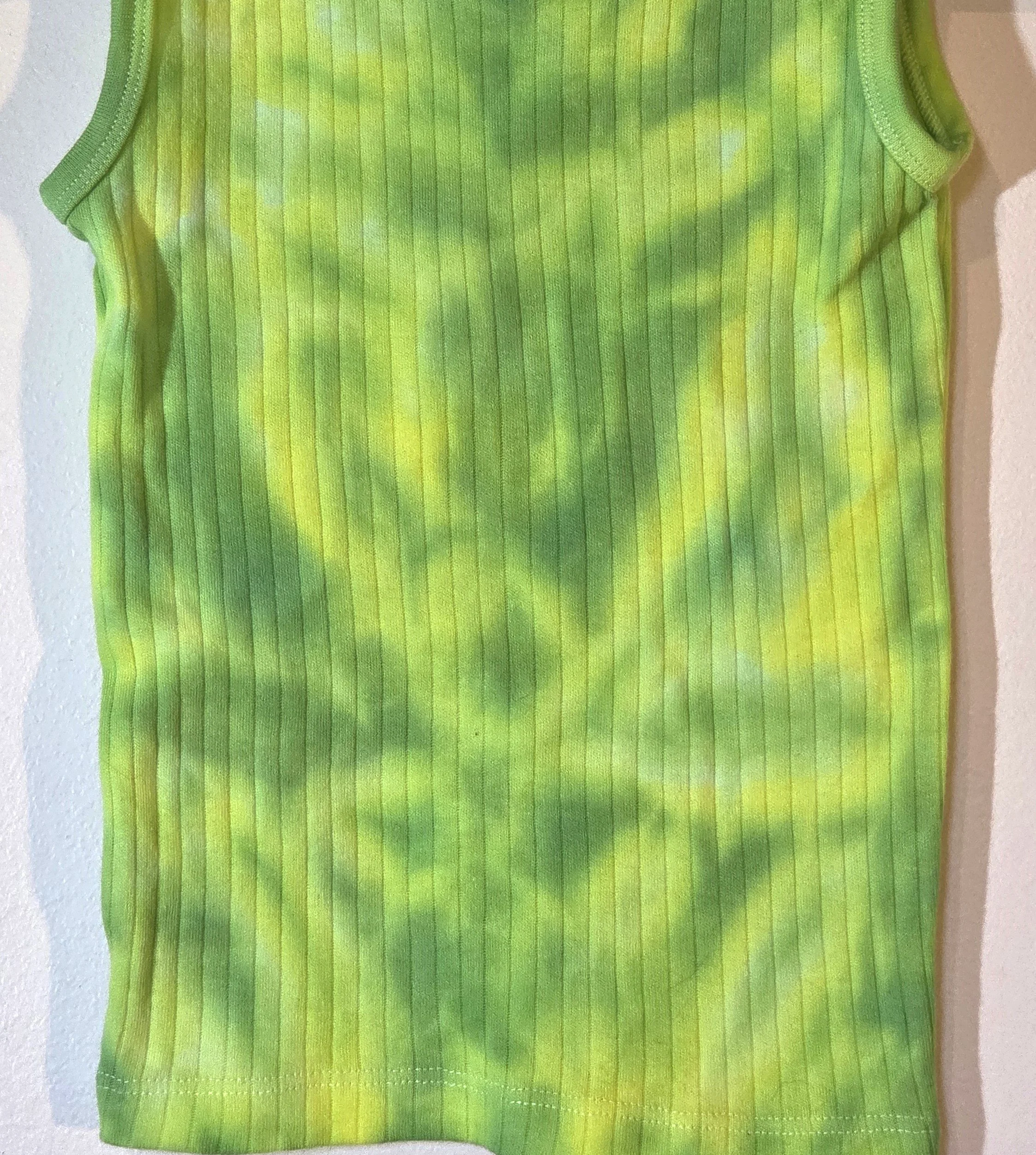 A child's ribbed singlet featuring a faint, pastel tie-dye pattern of lime green and yellow that creates a soft abstract flower shape near the bottom hem.