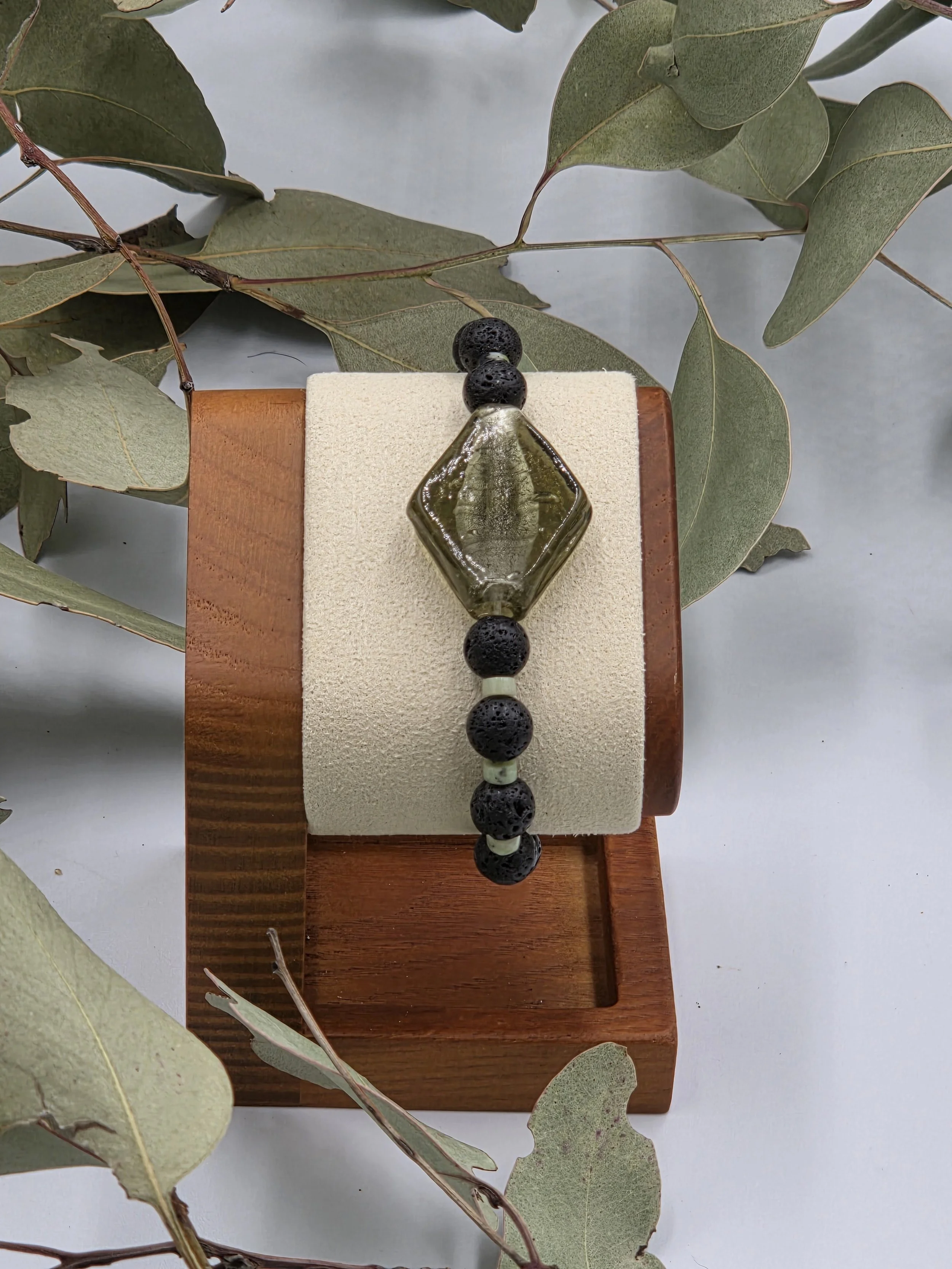 Olive green rhombus glass bead centered between alternating black lava beads and small green chrysoprase discs on an elastic bracelet.