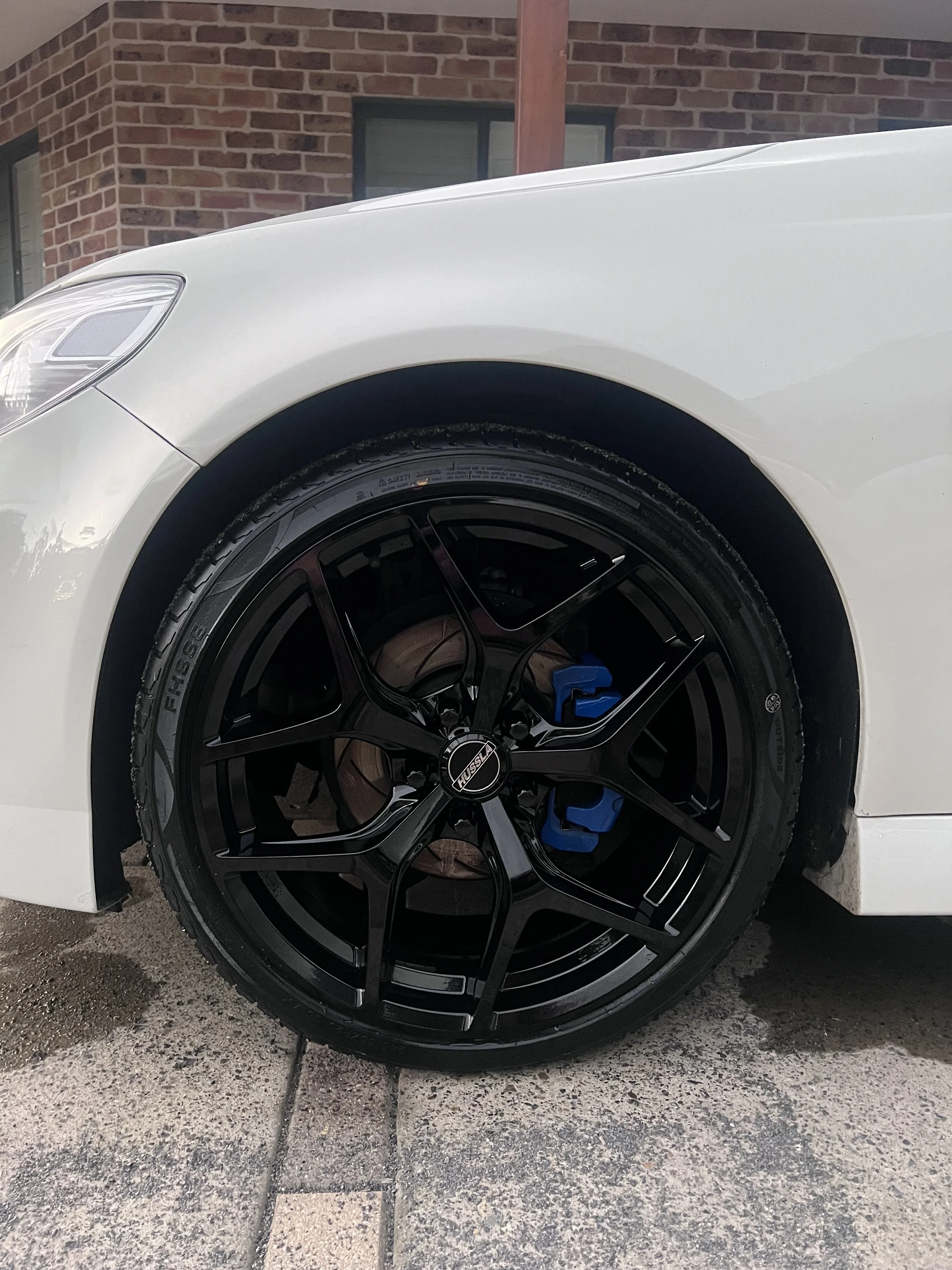 Close-up of a white car's front wheel with a black rim and blue brake caliper, parked on a concrete driveway near a brick building.