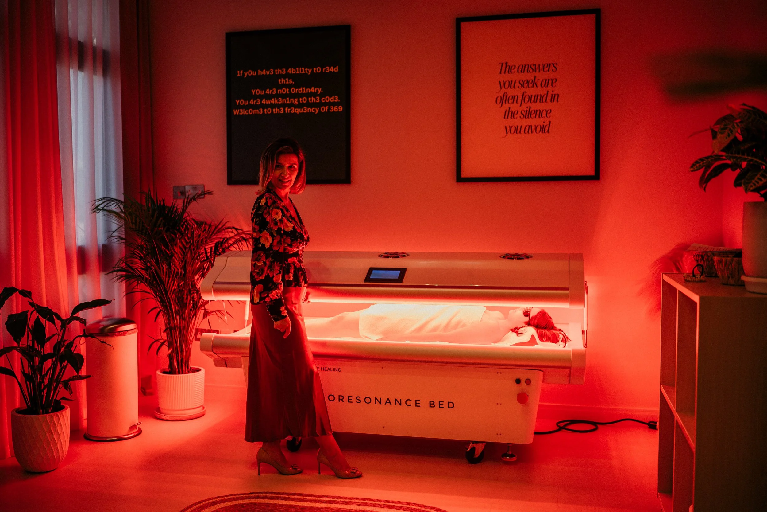 A woman standing in a dimly lit room with red lighting, next to a person lying inside a medical or therapy bed labeled "Resonance Bed." The woman is smiling and dressed in a floral top and long skirt, wearing high heels. The room has potted plants, framed artwork on the wall, and curtains on a window.