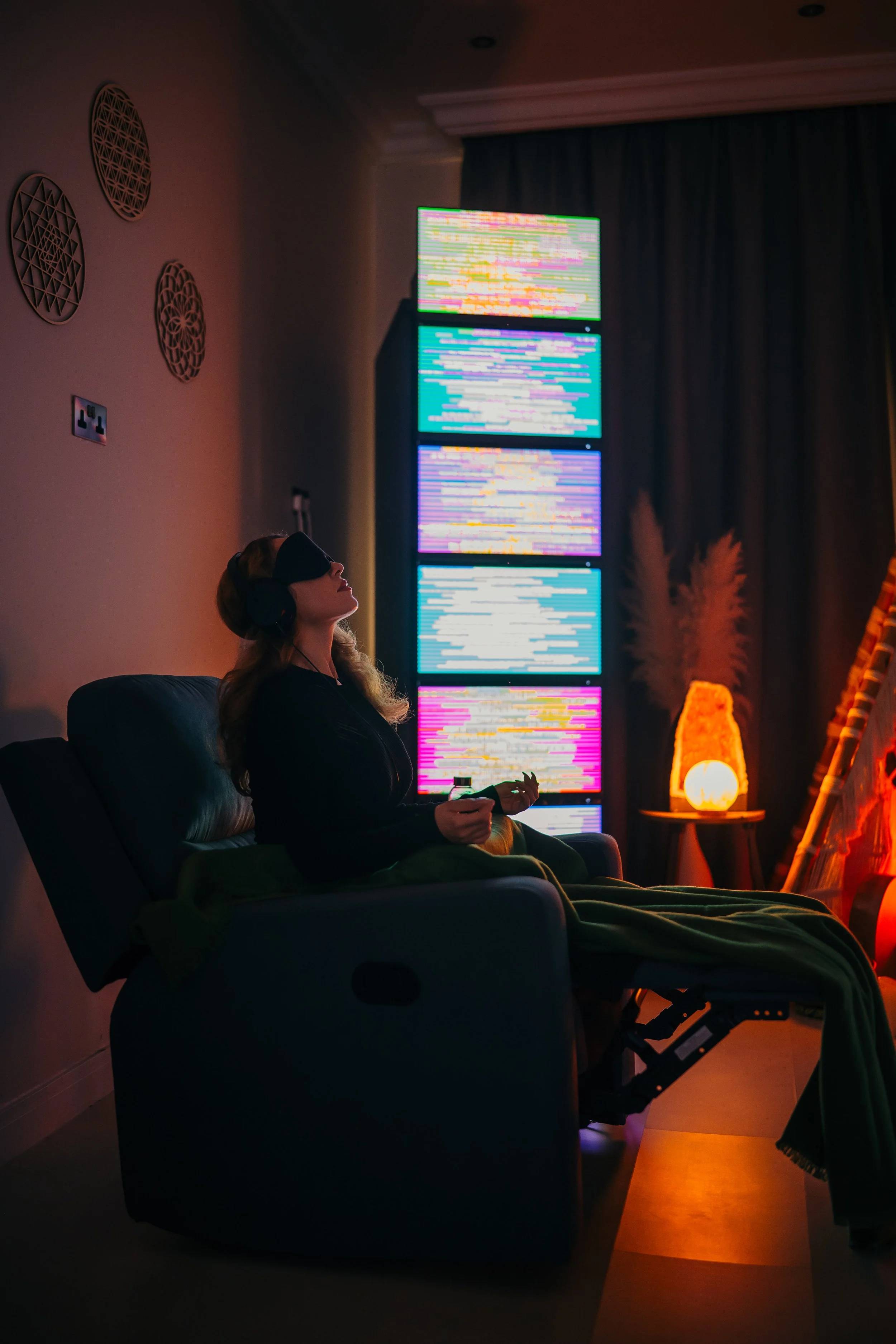 A woman wearing a virtual reality headset is sitting in a recliner, surrounded by colorful neon lights and digital screens in a dark room.