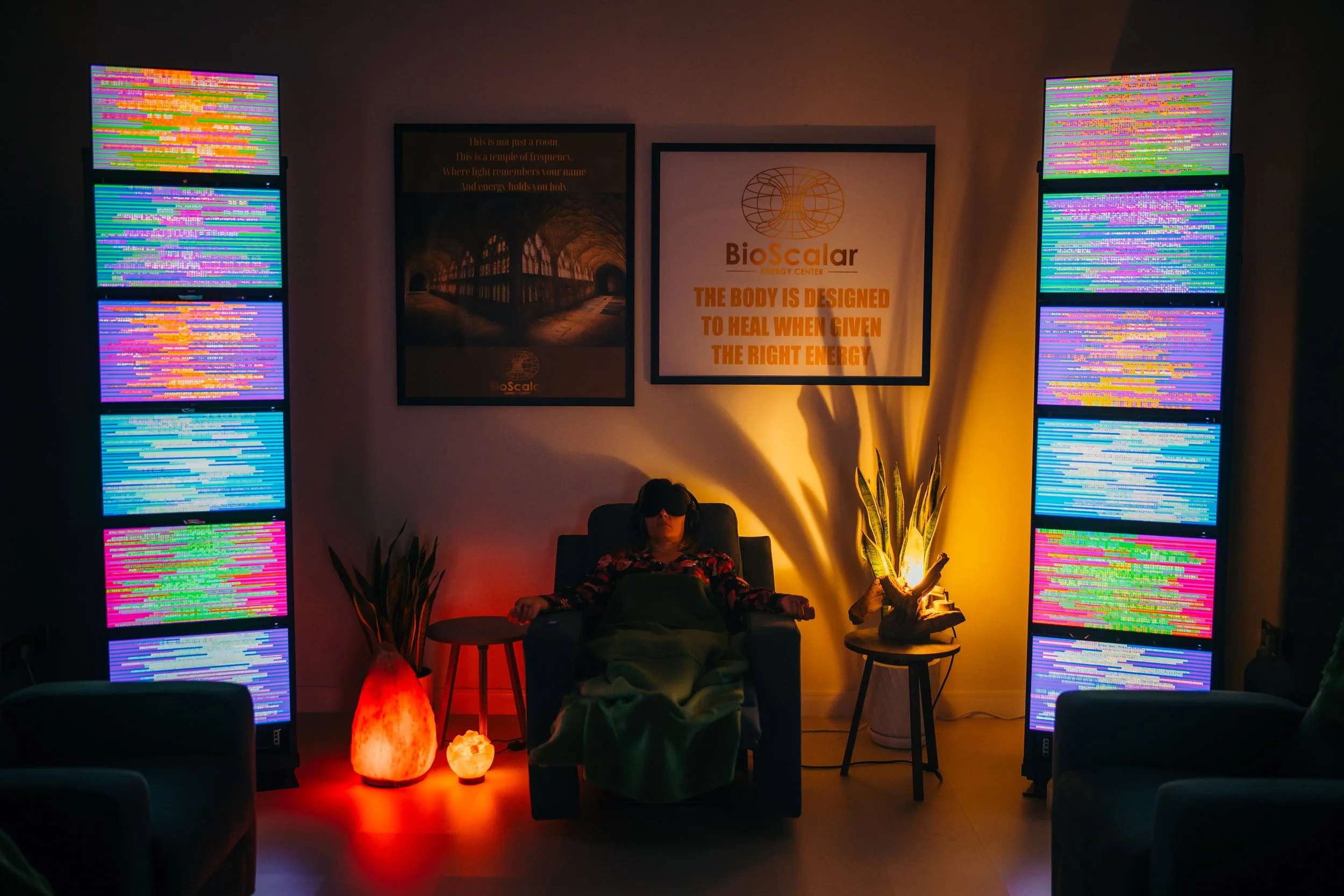 A woman in a chair with eye protection, surrounded by colorful LED light panels and ambient lighting, in a wellness or relaxation space with rollers and plants.