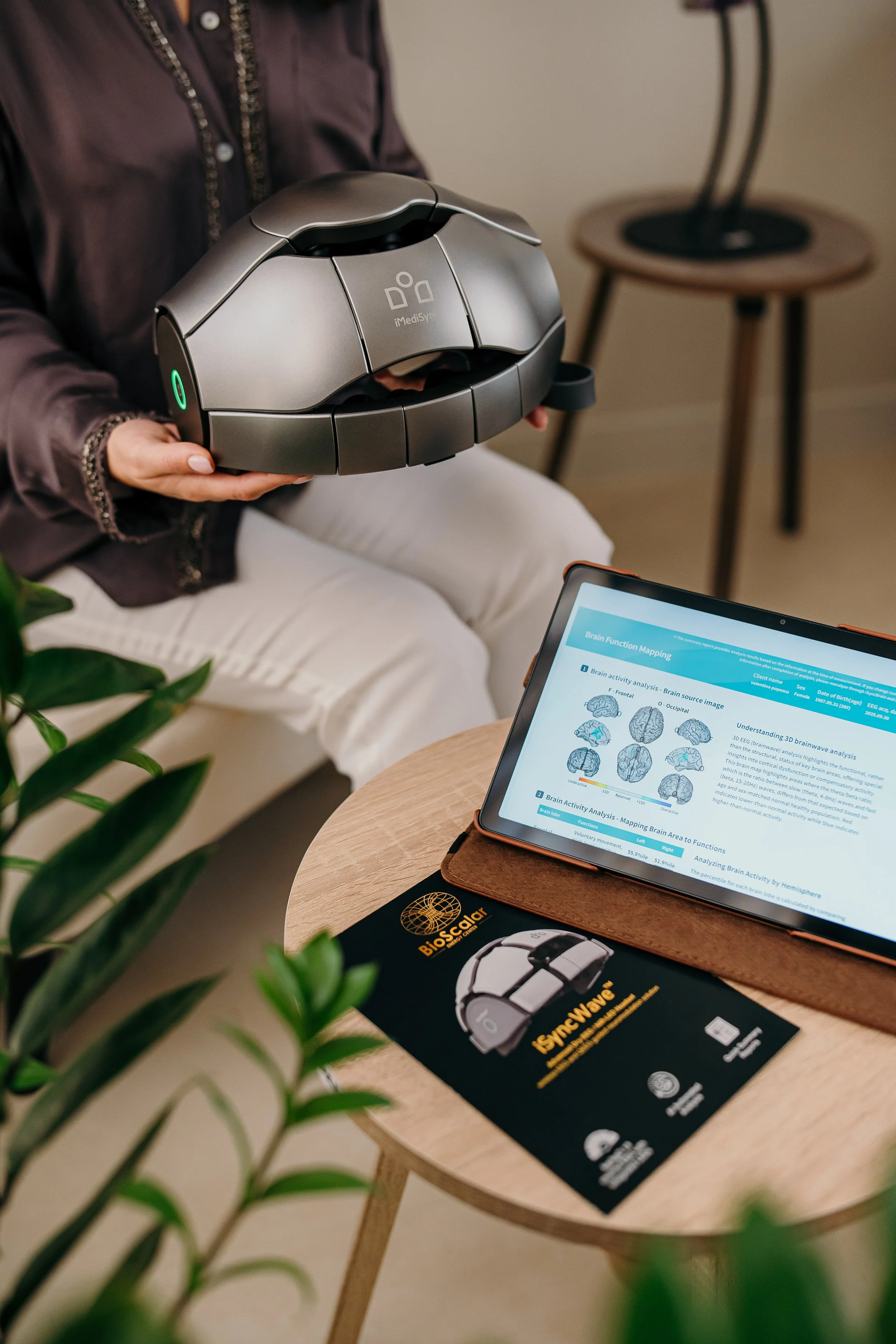 A person holding a modern medical or diagnostic device called iMediSYRE, with a tablet displaying brain function analysis and mapping. The device and tablet are on a wooden table, with a brochure for a BioSensor and SyncWave placed nearby. The setting appears to be a clinic or medical office.