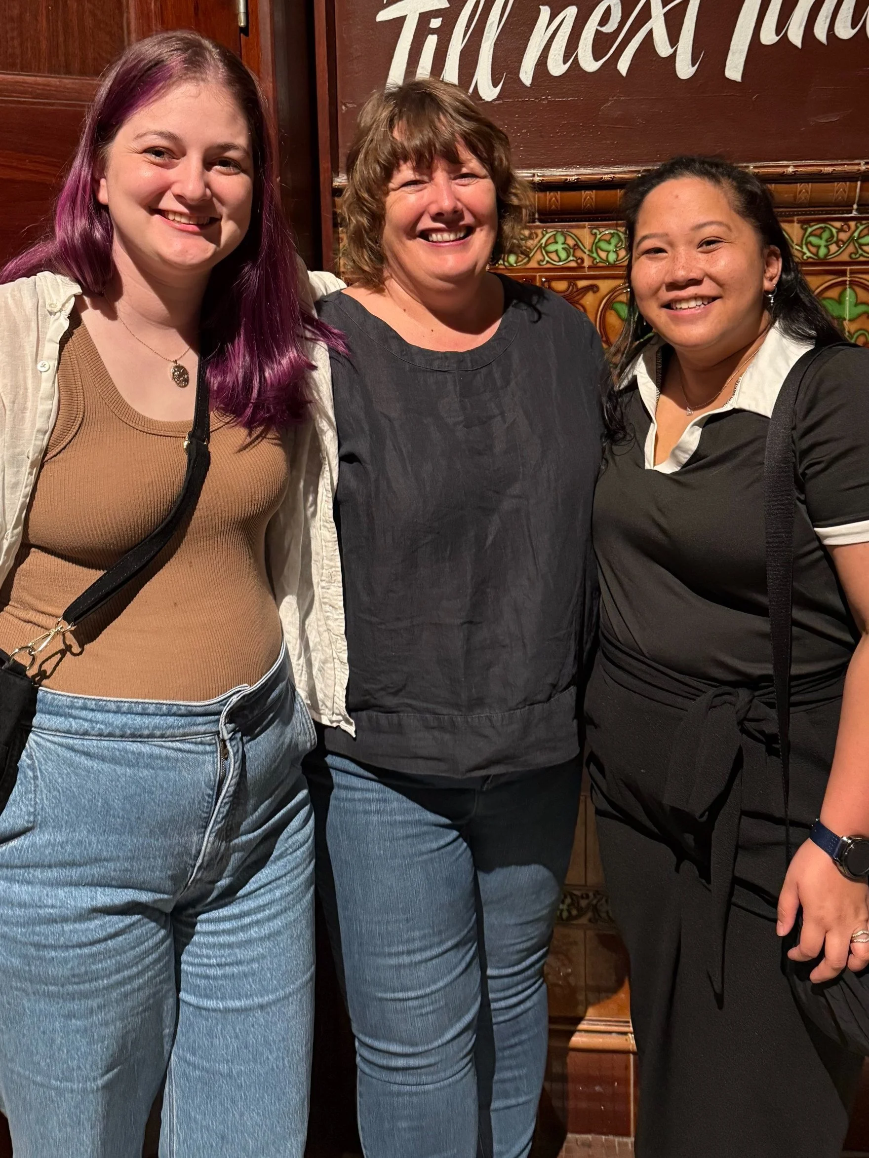 Three women smiling and standing close together in front of a wooden wall with writing and decorative tiling, in a warm, casual setting.