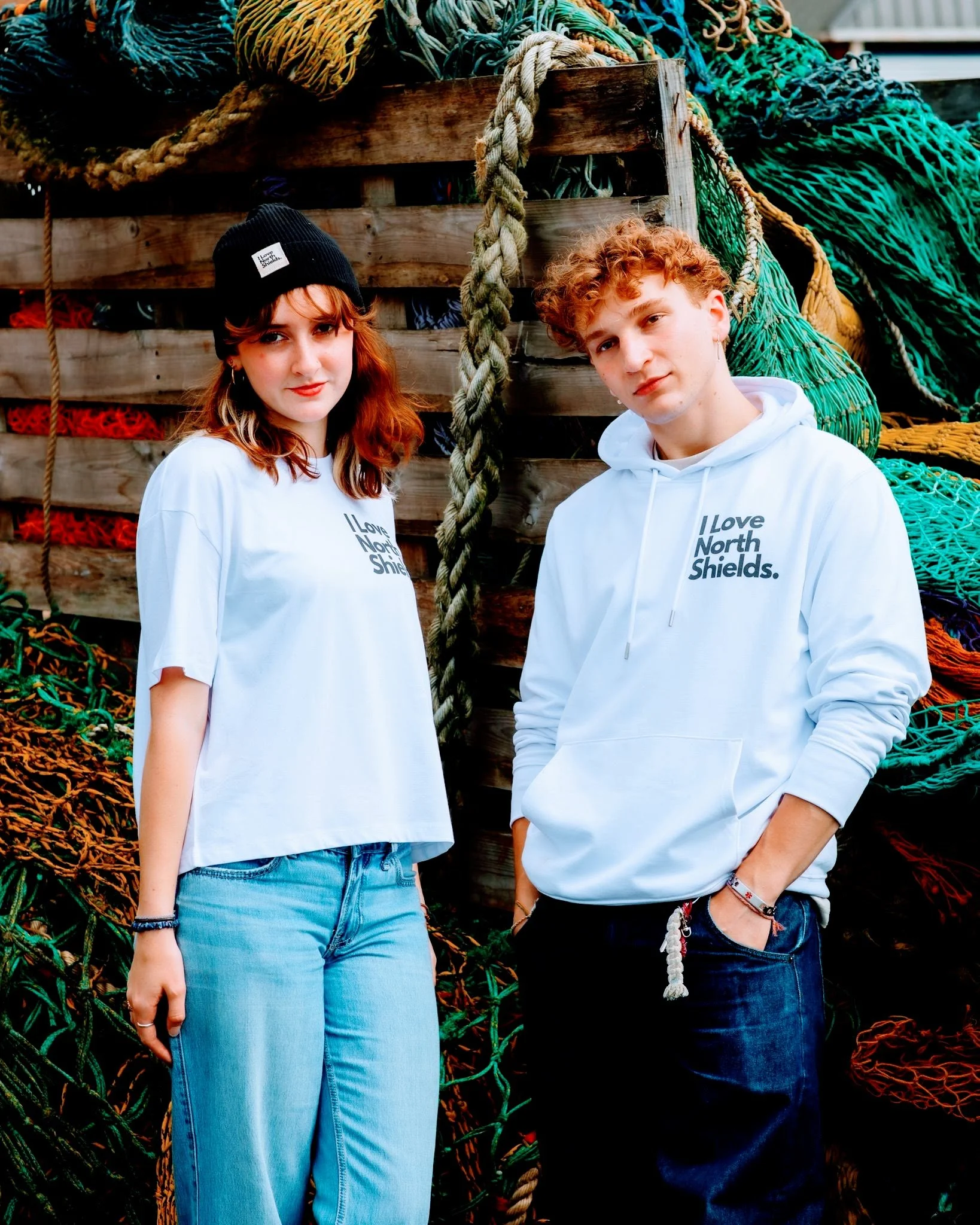 Two young people, a woman with red hair wearing a black beanie and a white t-shirt, and a man with curly red hair in a white hoodie, standing outdoors against a background of colorful fishing nets and ropes.