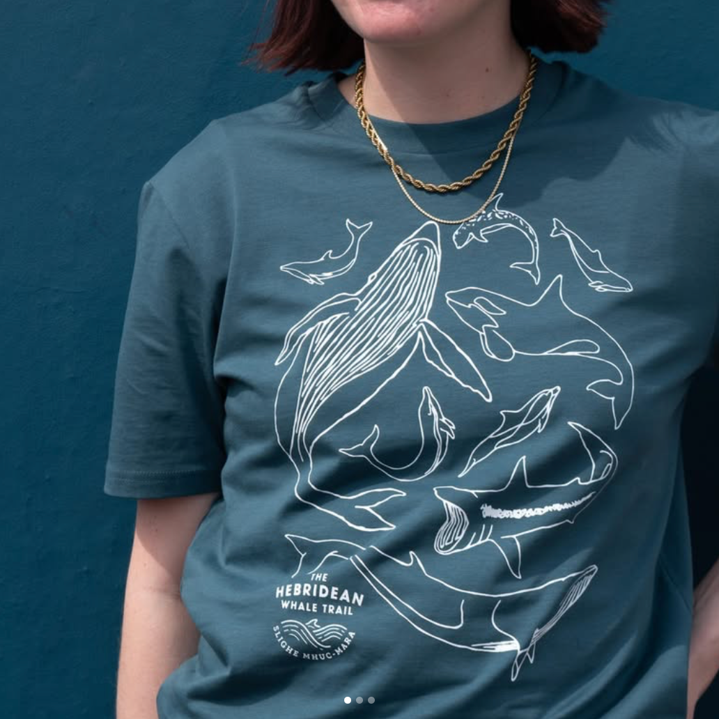 Close-up of a teal 100% organic cotton t-shirt with white outline illustrations of various whales and dolphins, and a small logo that reads 'The Hebridean Whale Trail.' The t-shirt was printed by Lyoness Press for a charity merchandise collection.