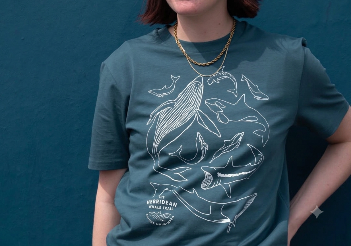 A woman wearing a teal T-shirt with a white line drawing of various whale species and a text that reads "The Hebridean Whale Trail." She is also wearing a gold chain necklace and standing against a matching teal background.