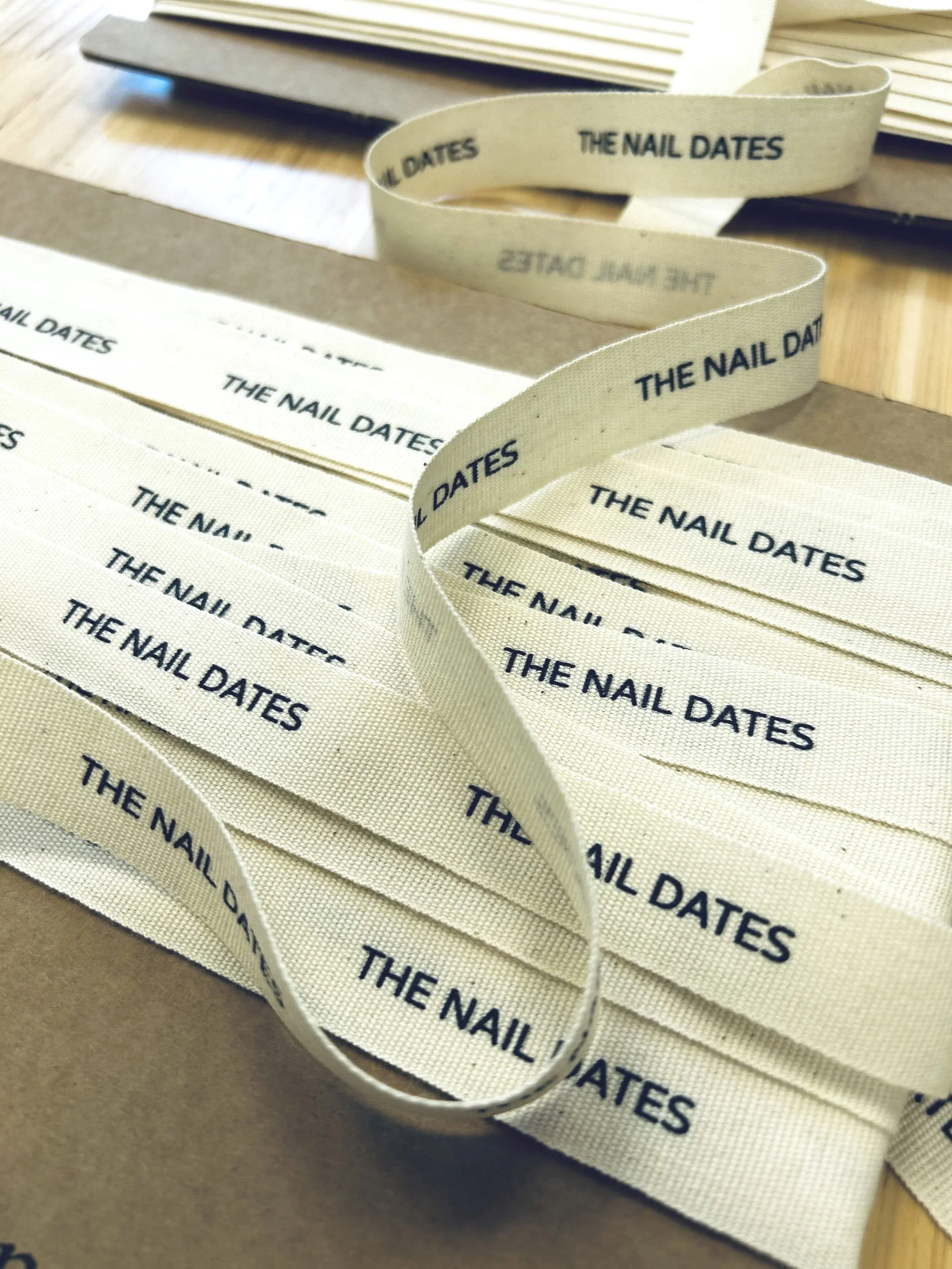 Close-up of cream-colored ribbon with black text that reads 'The Nail Dates,' laid on wooden and paper surfaces, with additional ribbons and papers in the background.