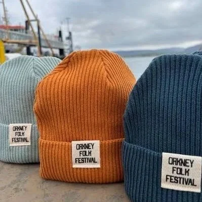 Three colourful 100% organic cotton beanies, each with a printed cuff which says "Orkney Folk Festival". The beanie cuffs are made from 100% cotton and are printed with eco-friendly inks at the Lyoness Press studio.