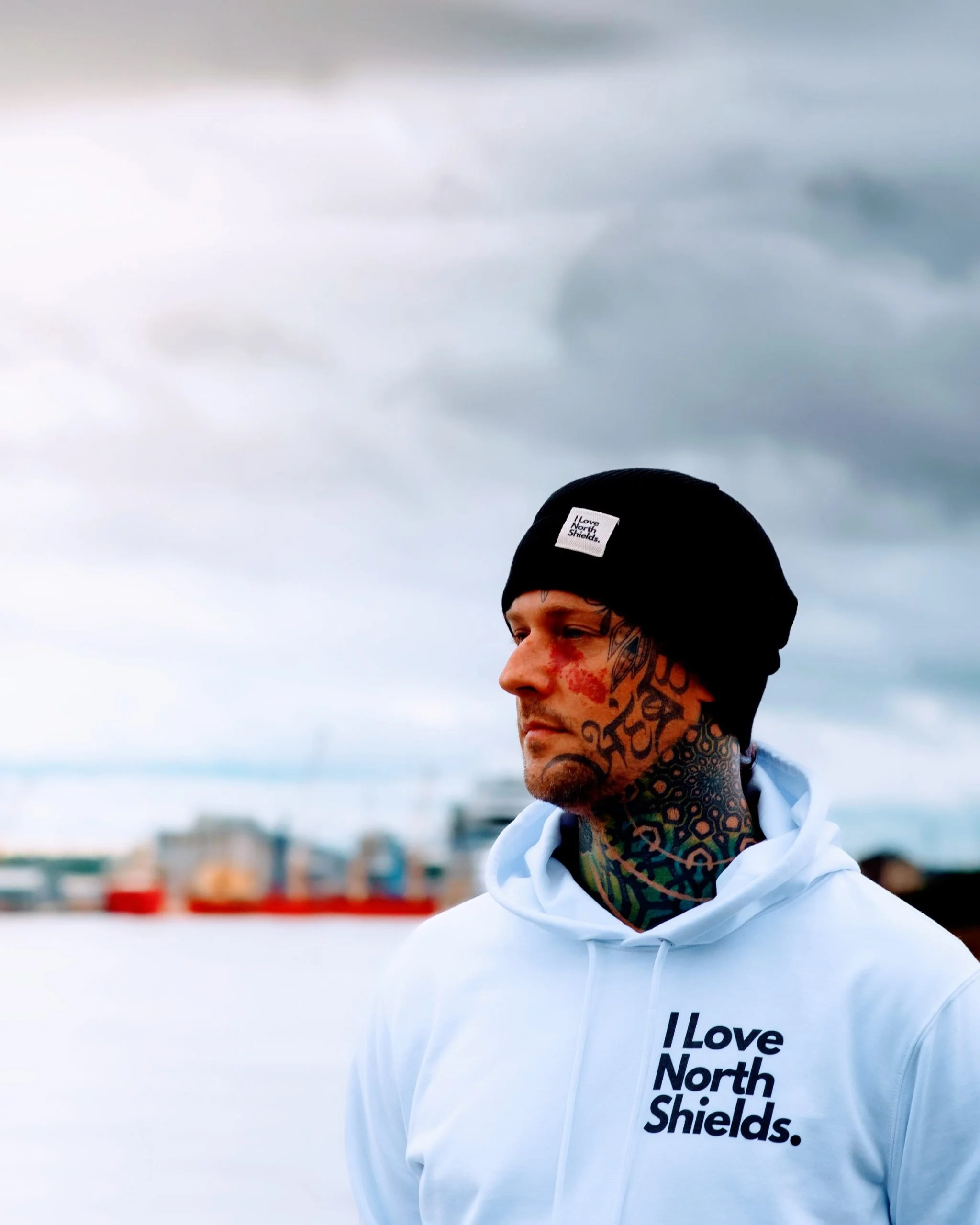 A man with tattoos on his face and neck, wearing a black beanie with a label and a white hoodie that has text 'I Love North Shields,' standing outdoors against a cloudy sky.