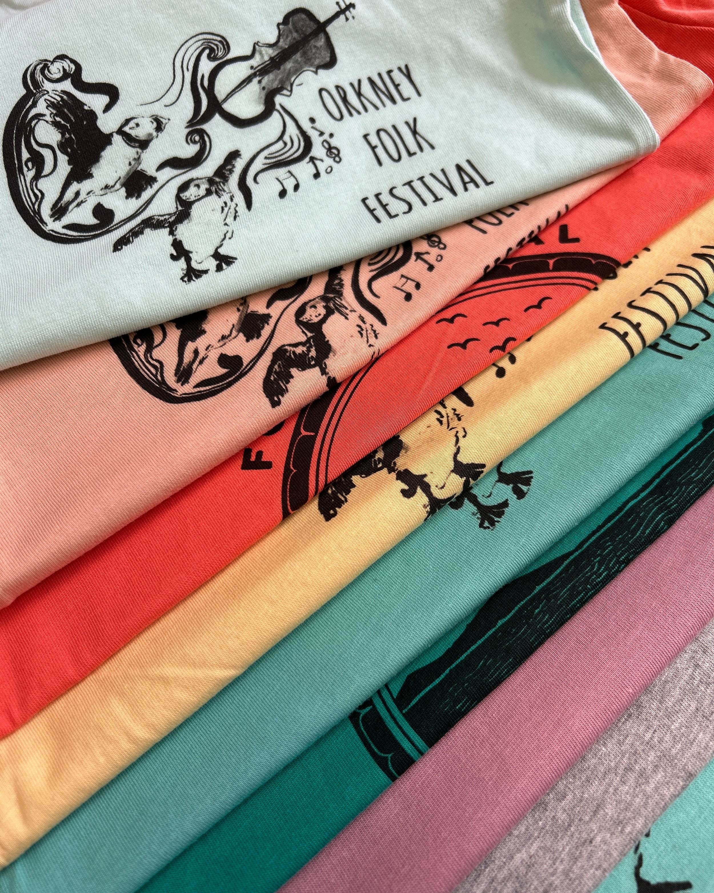 Stack of colorful T-shirts with a design and text related to the Oakney Folk Festival.