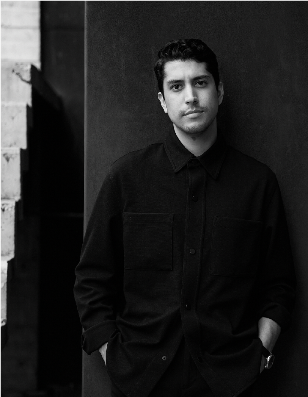 Black and white photo of a young man with dark hair, wearing a dark button-up shirt, standing with his hands in his pockets against a wall.