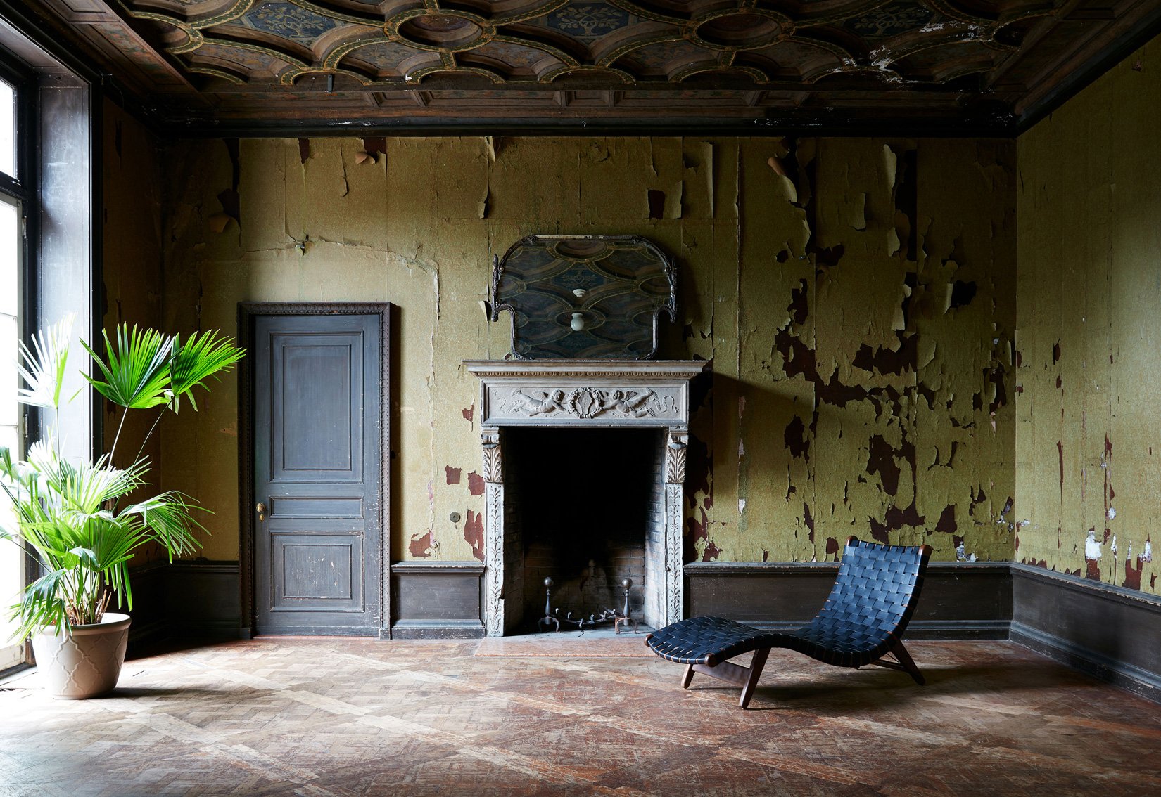 An old, abandoned room with peeling yellow wallpaper, a fireplace with an ornate mantel, a black door, a green potted plant by a large window, and a black woven lounge chair.
