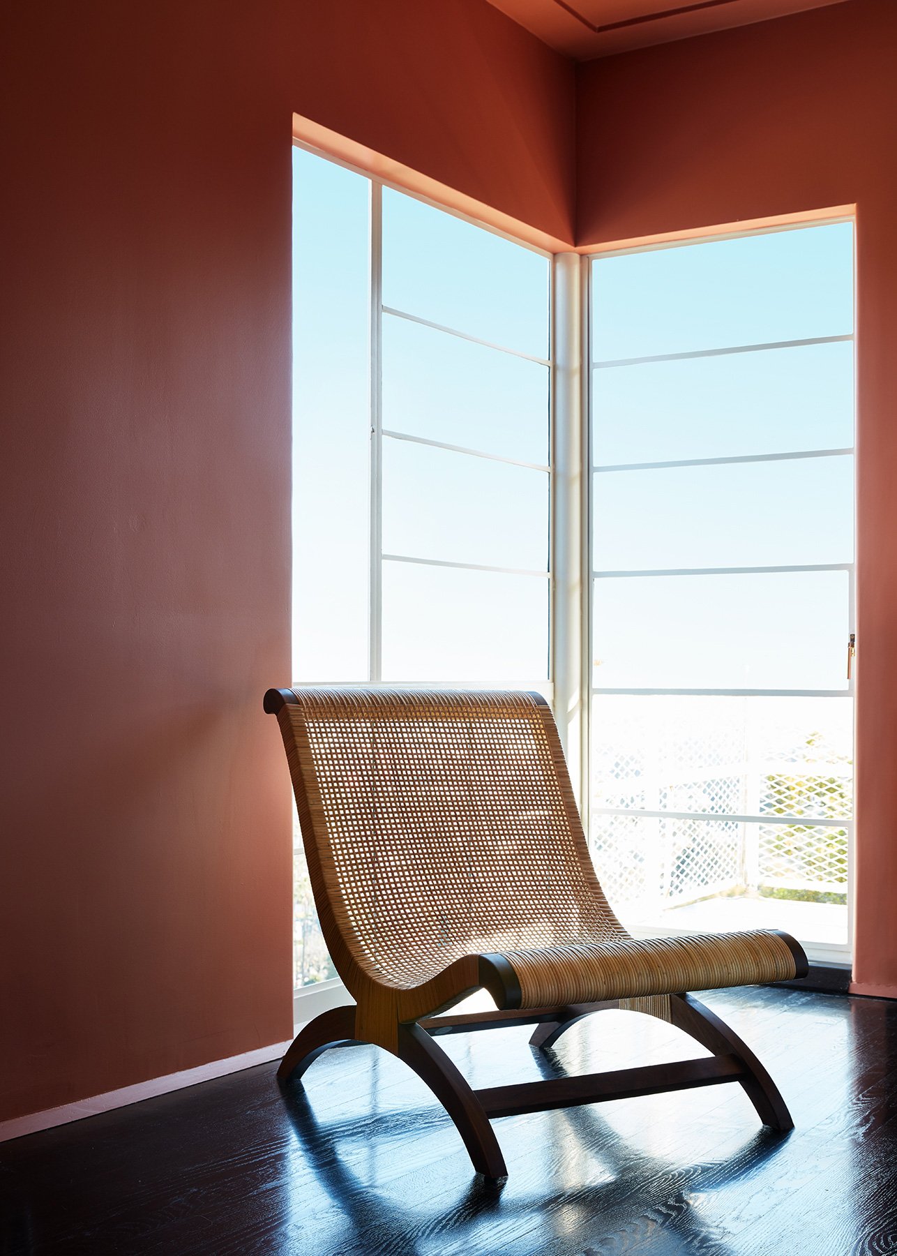 A modern interior scene with a wooden lounge chair featuring a curved woven rattan seat and backrest, positioned in front of a large corner window with multiple panes, letting in bright sunlight.