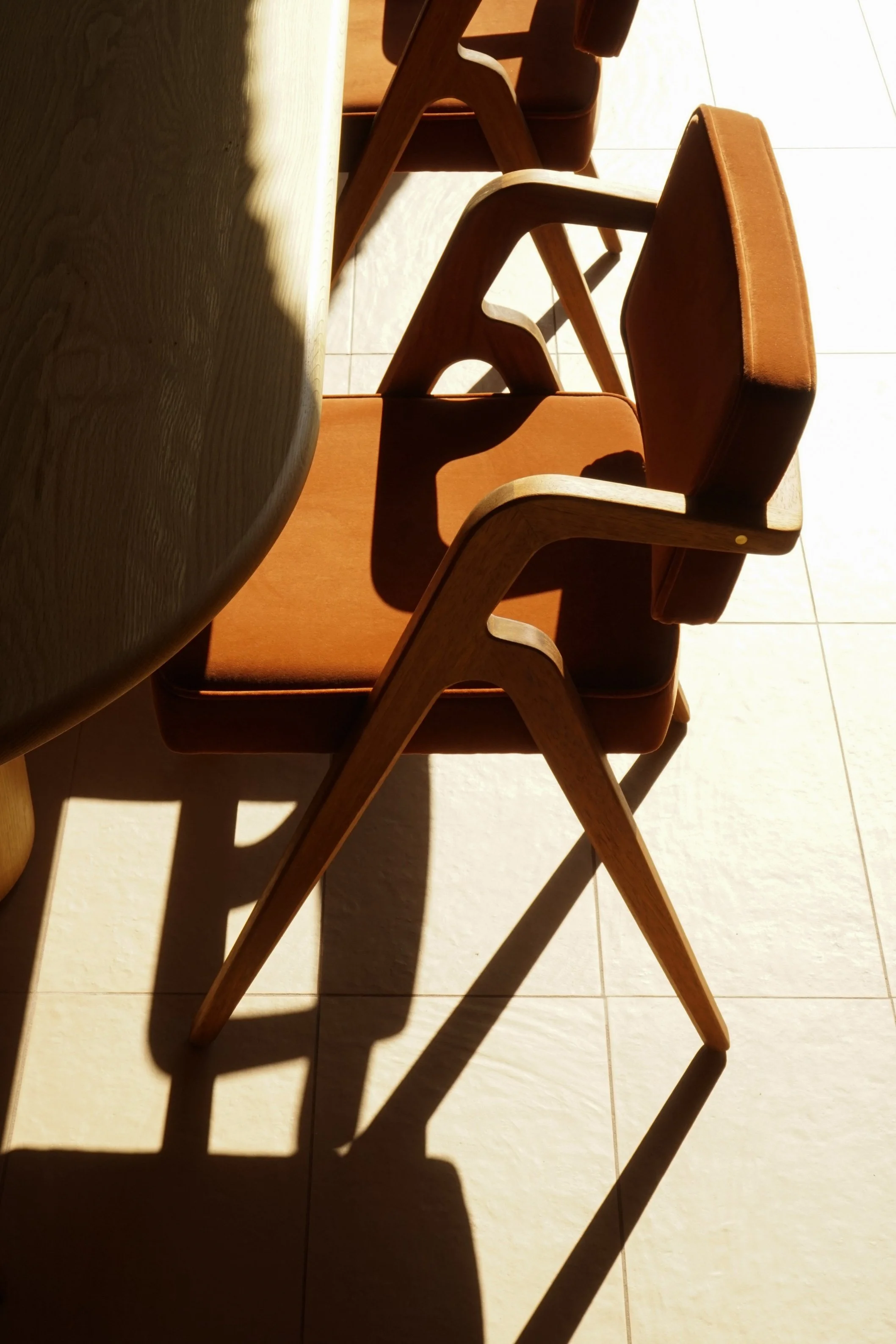 A wooden dining table with four chairs around it, partially in sunlight, casting shadows on a tiled floor.
