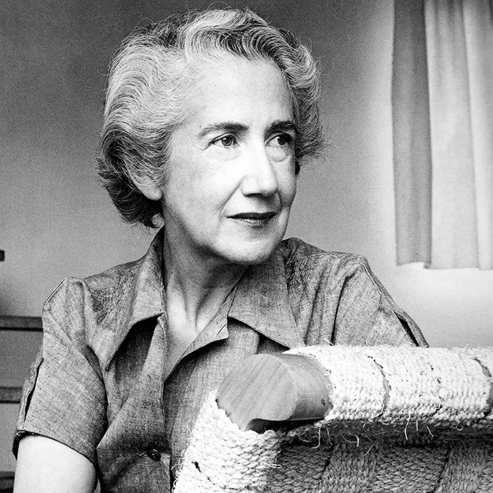 Black and white portrait of an elderly woman with short, wavy hair, wearing a collared shirt, sitting with her arms resting on a woven armrest.