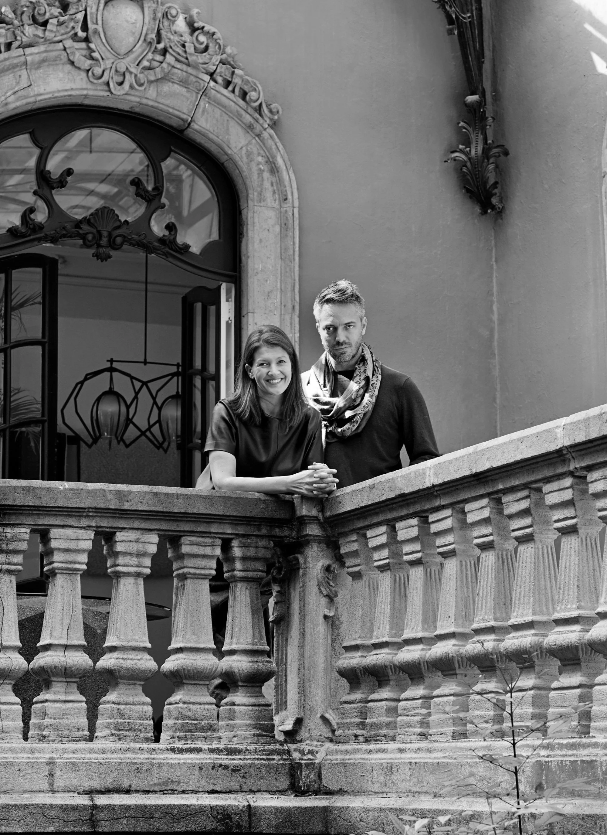 Black and white photo of a smiling woman and a serious man standing on a balcony with ornate stone railings, behind an open window with decorative ironwork, in what appears to be a historic building.