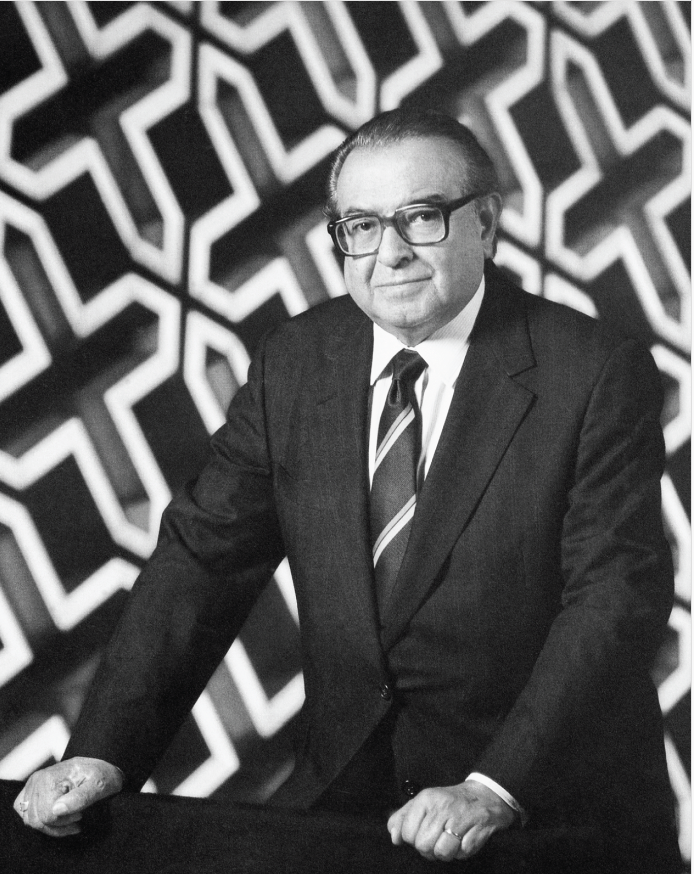Black and white photograph of a middle-aged man with glasses in a suit and tie, standing with hands on a table, against a geometric pattern background.