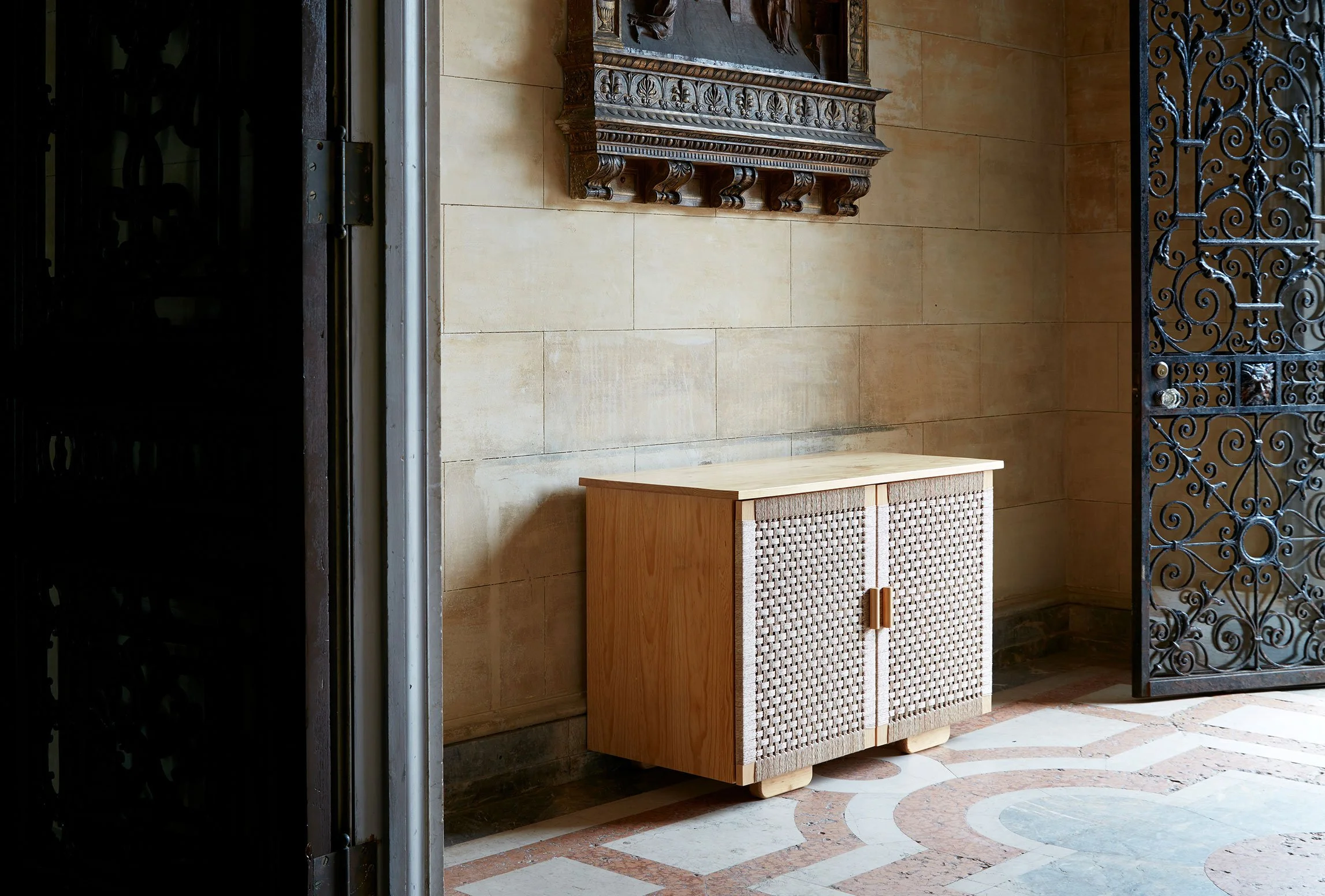 A wooden cabinet with woven doors is positioned against a beige tiled wall, next to a black ornate iron gate, in a room with a patterned tile floor.