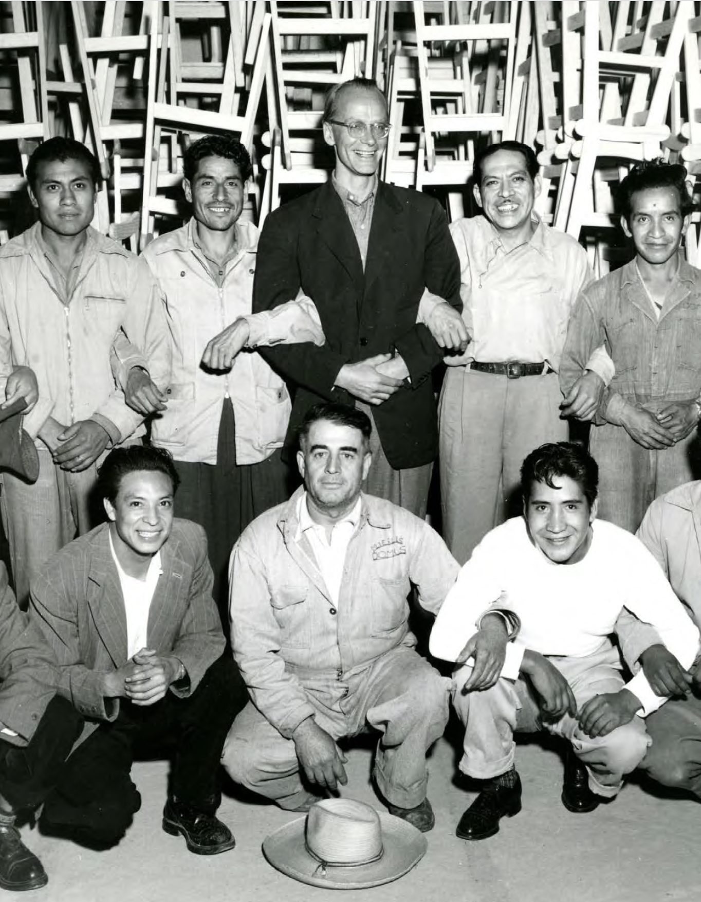 A group of nine men, most wearing work clothes, posed indoors with chairs stacked in the background. The man in the center of the back row is taller and wearing glasses. A hat is on the ground in front of the group.
