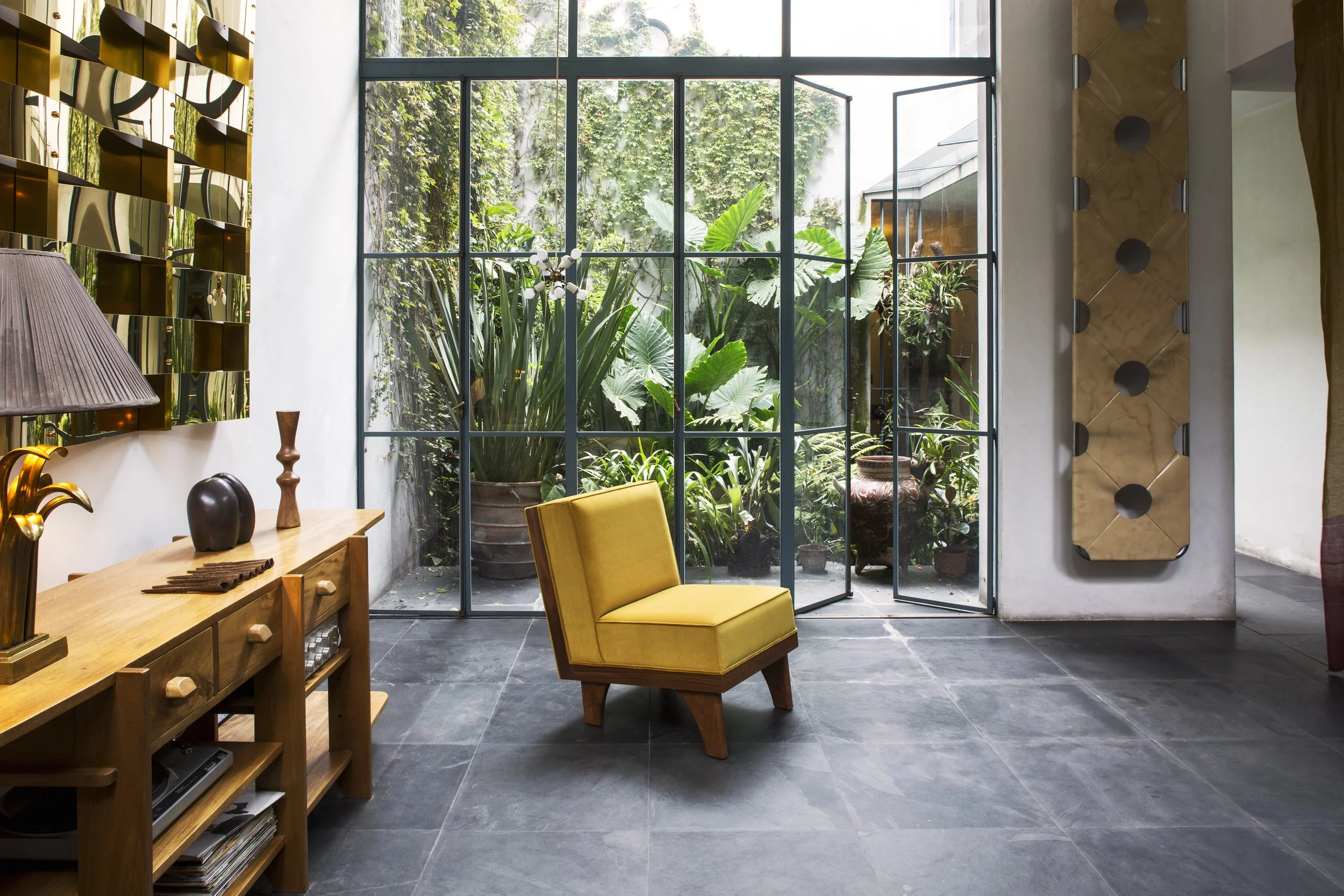 Bright living room with large glass doors opening to a lush outdoor garden, featuring a yellow chair, wooden console table with decorative objects, and modern wall decor.