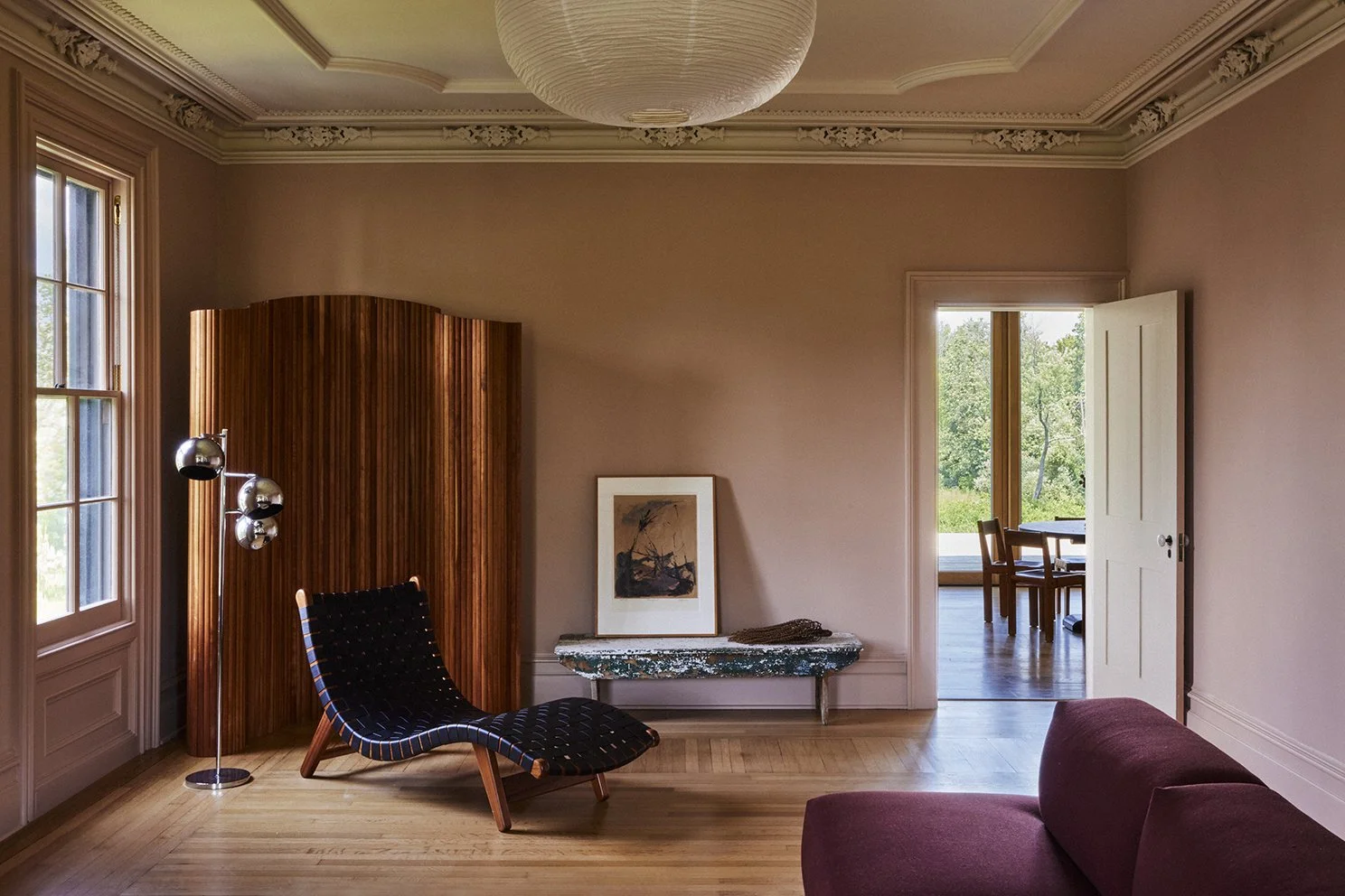 Interior of a living room with pink walls, a wooden floor, a black lounge chair, a tall multi-bulb floor lamp, a framed artwork leaning against the wall, a marble-topped side table, an open door leading to a bright dining area, and large windows letting in natural light.