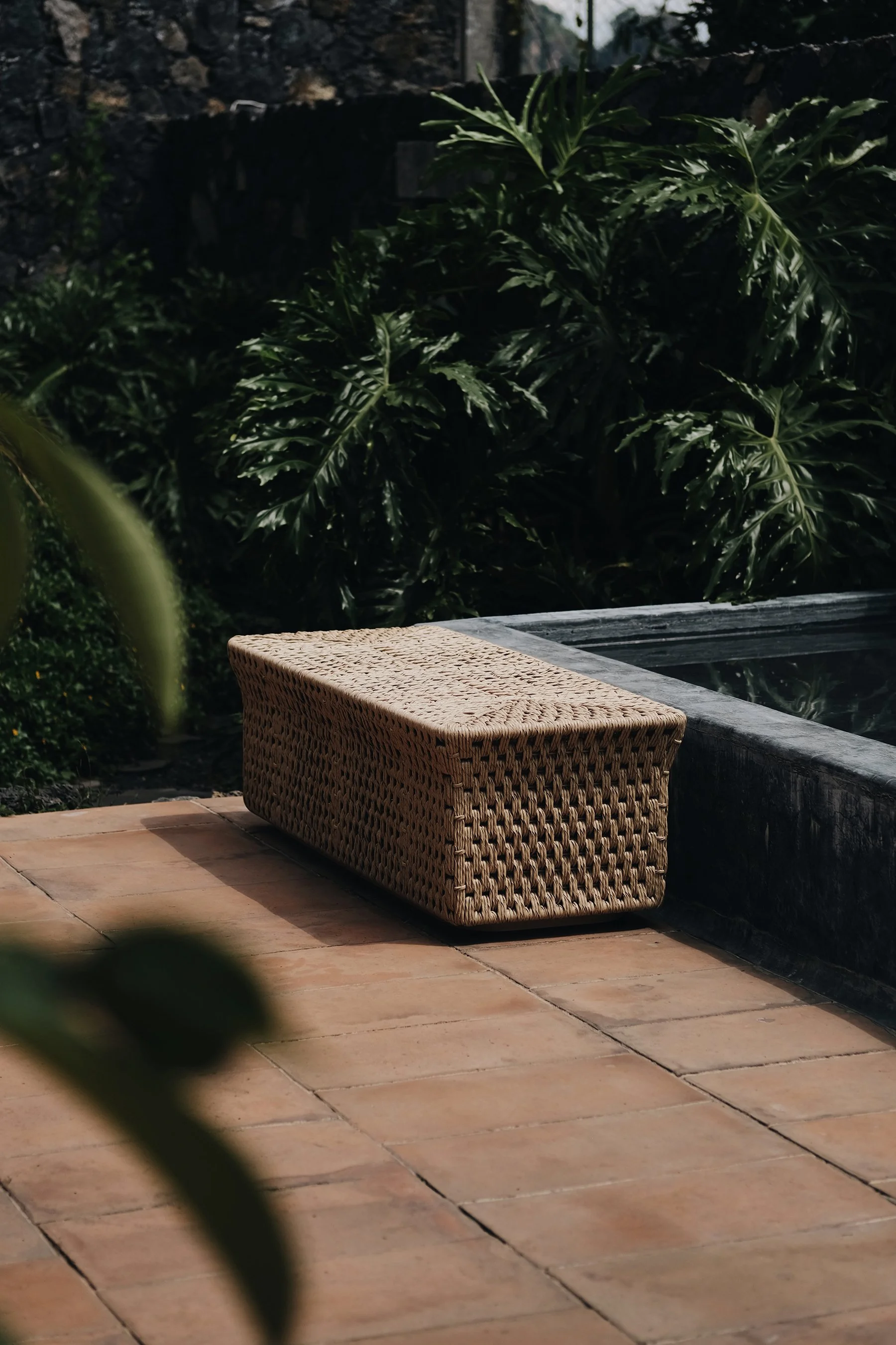 Wicker outdoor bench on a tiled patio near a pool, with lush green plants and bushes in the background.